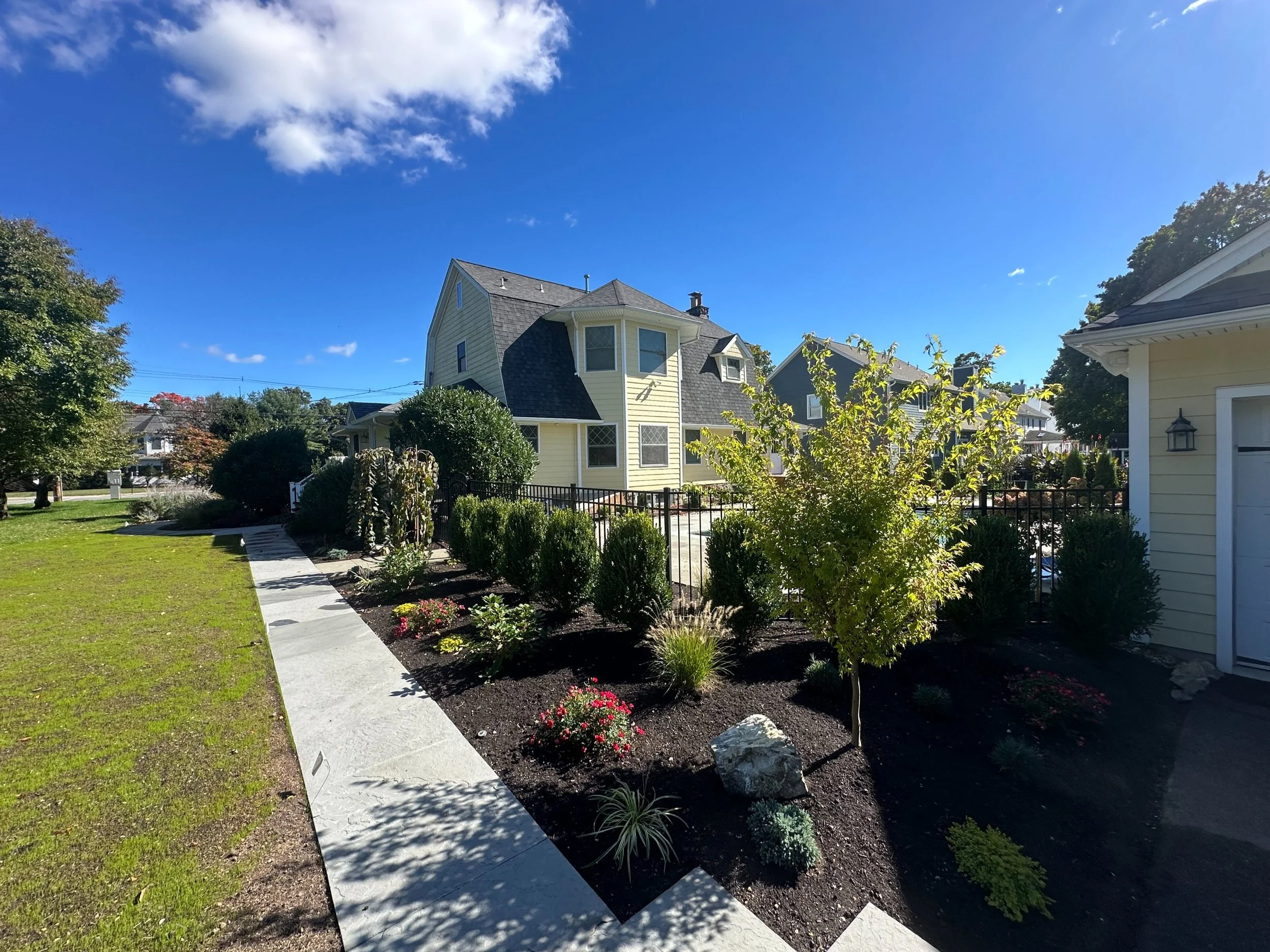A residential neighborhood with houses, a well-maintained lawn, a sidewalk, trees, and bushes under a bright blue sky with some white clouds.