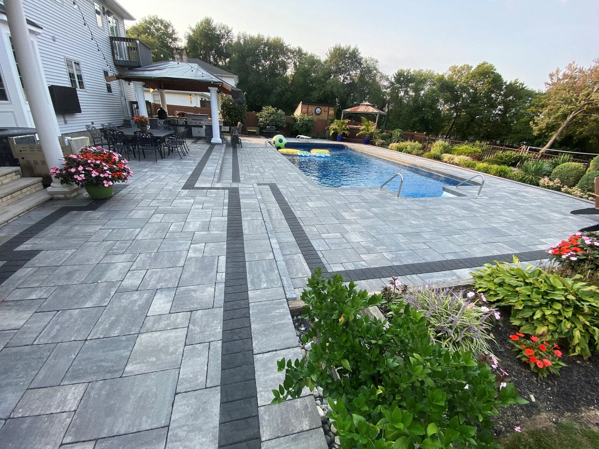 A backyard patio with a swimming pool surrounded by a landscape of green trees and bushes, with outdoor furniture including a table with chairs, umbrellas, a grill area, and colorful potted plants and flowers.