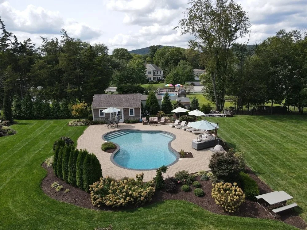 A backyard with a kidney-shaped swimming pool, surrounded by a paved patio area with lounge chairs and umbrellas, lush green grass, garden beds with bushes and flowers, and a small pool house with seating. In the background, there are trees, a fence,
