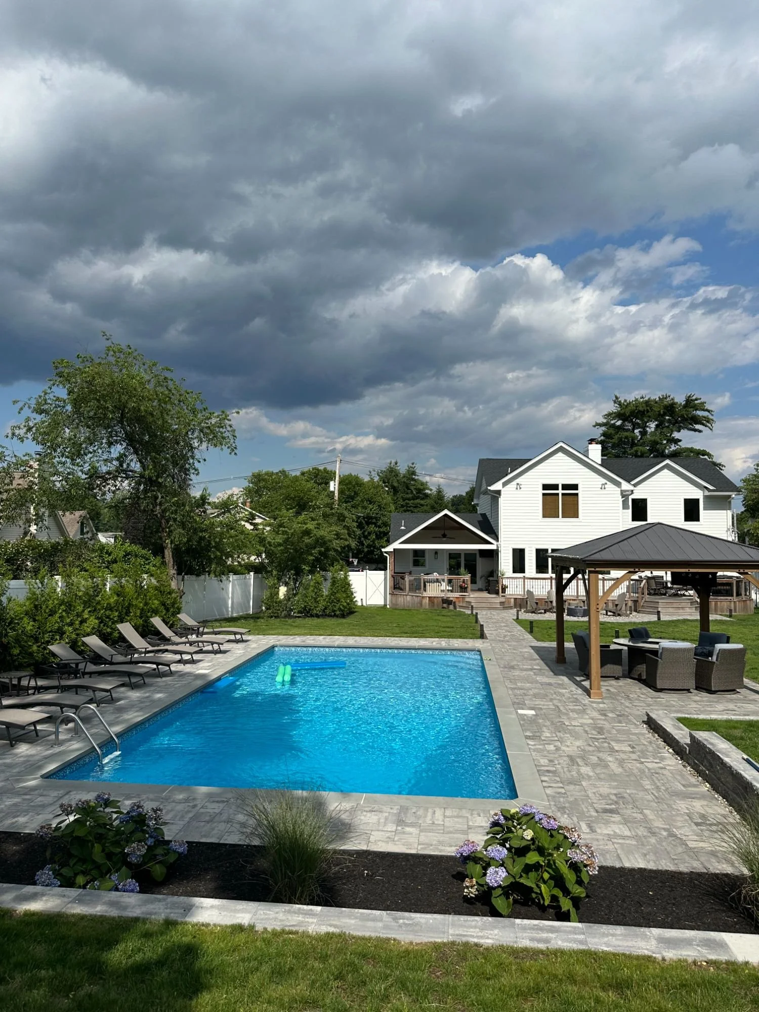 Rectangular pool with paver surround, lounge chairs, and a pergola in a landscaped residential backyard — Green Nature Landscape Design