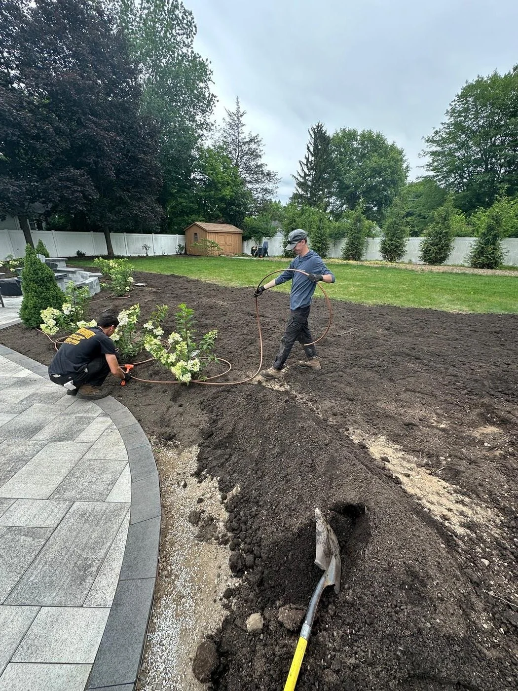 Two landscapers installing plantings and irrigation tubing in a freshly graded backyard alongside a curved paver patio. — Green Nature Landscape Design
