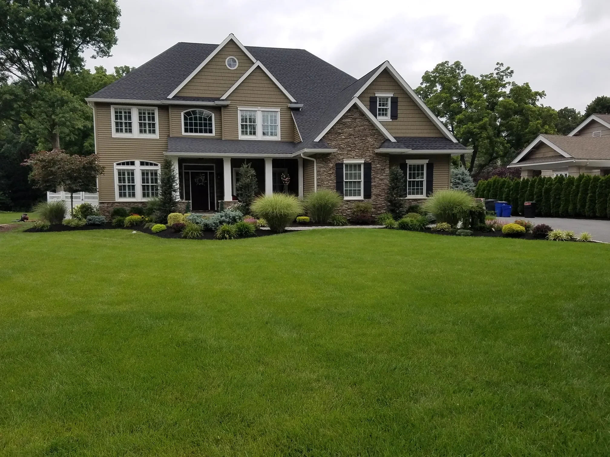 Completed front yard landscape design with layered shrubs, ornamental grasses, and a manicured lawn in front of a large two-story home. — Green Nature Landscape Design