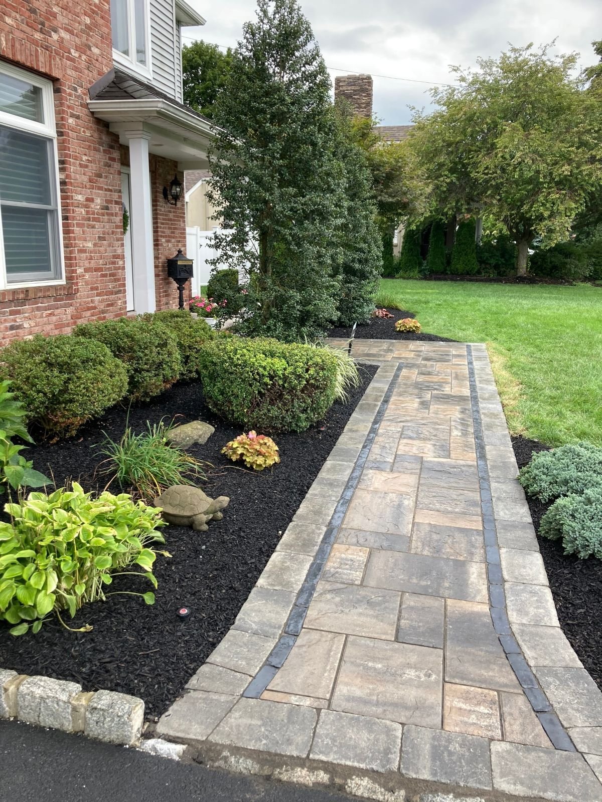 Paver walkway with dark border edging leading to a brick home entrance surrounded by mature shrubs, hostas, and black mulch beds. — Green Nature Landscape Design