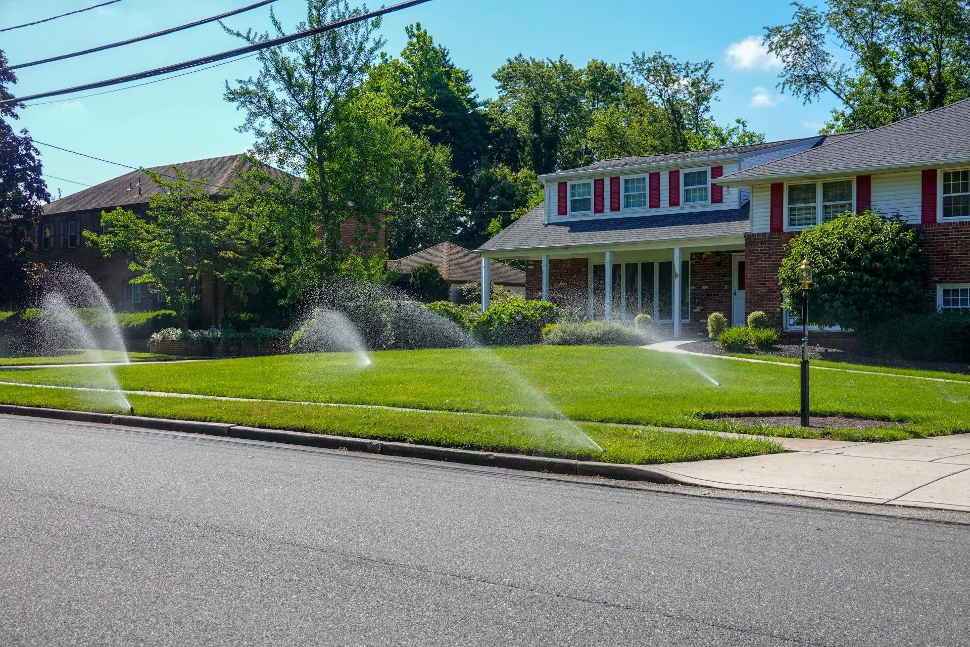 Sprinkler system watering a green front lawn at a two-story brick and siding home on a sunny residential street.