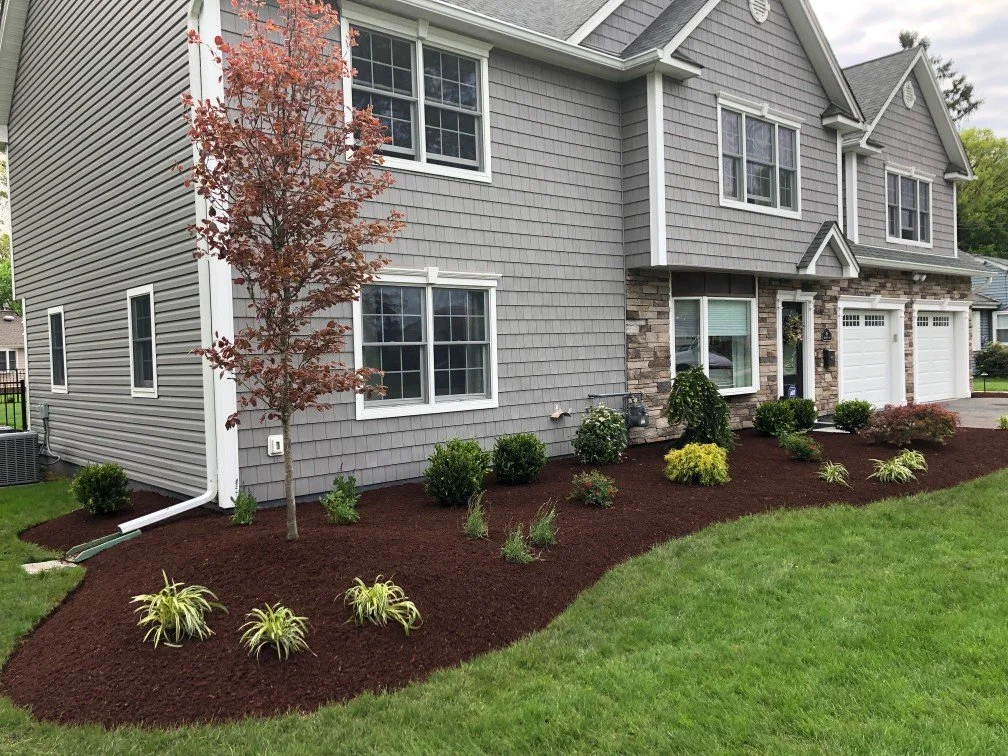Front yard landscaping with a young tree, shrubs, and flowering plants in front of a gray house with white trim and attached garage.