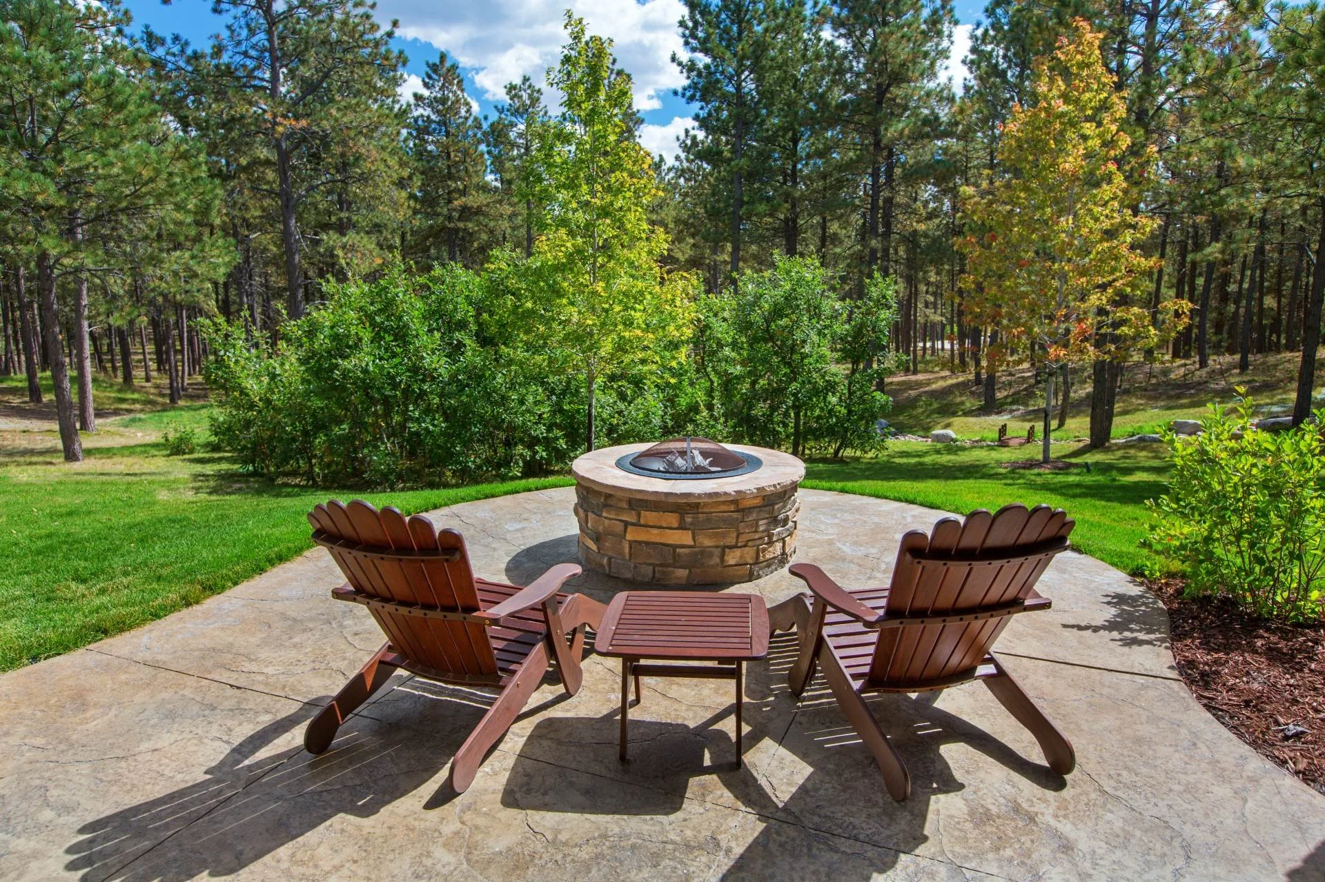 Round stone fire pit with Adirondack chairs on a stamped concrete patio surrounded by mature trees and green lawn