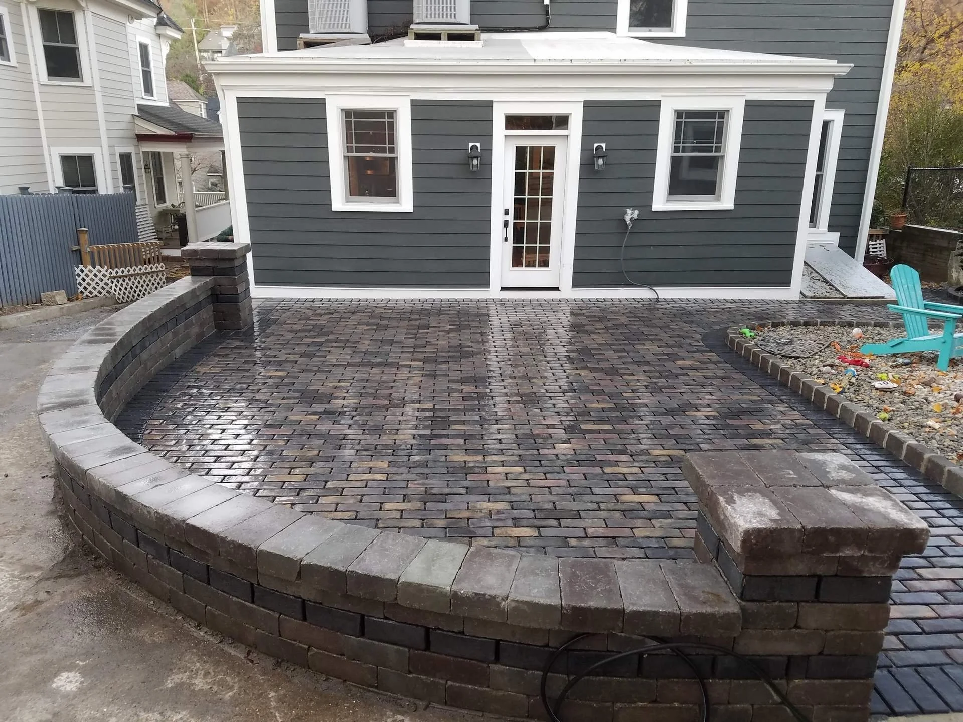 A newly paved brick patio with a curved brick border in front of a gray house with white trims and three windows, with a small garden area to the right and neighboring houses visible in the background.