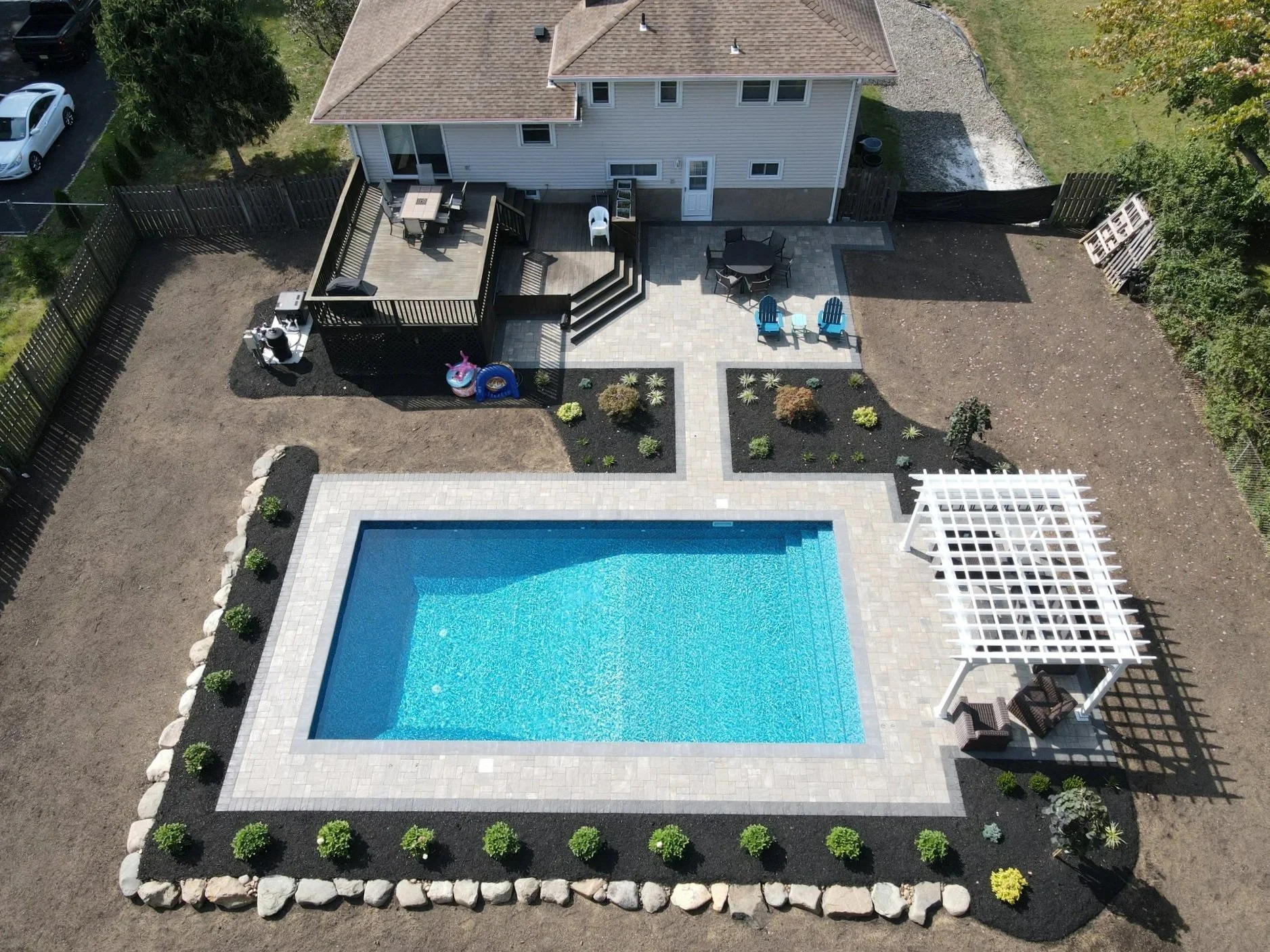 A backyard with a swimming pool, a wooden deck, outdoor furniture, and landscaping, surrounded by a wooden fence.