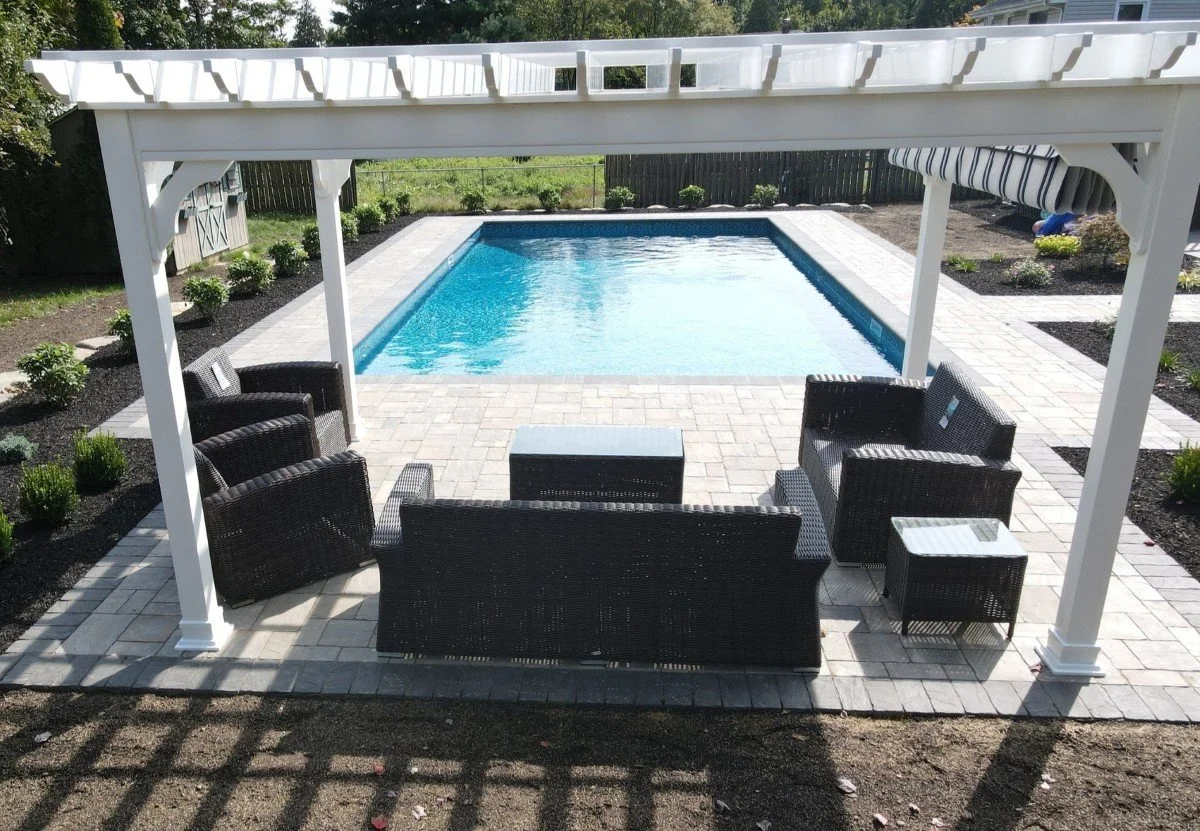 A backyard patio with a swimming pool, outdoor couches, and a pergola overhead, with landscaping and a garden surrounding the area.