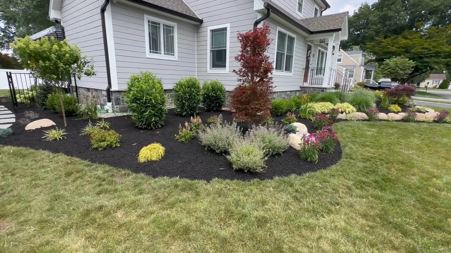 Curved landscape bed with mixed shrubs, ornamental grasses, and seasonal plantings against a dark mulch backdrop — Green Nature Landscape Design
