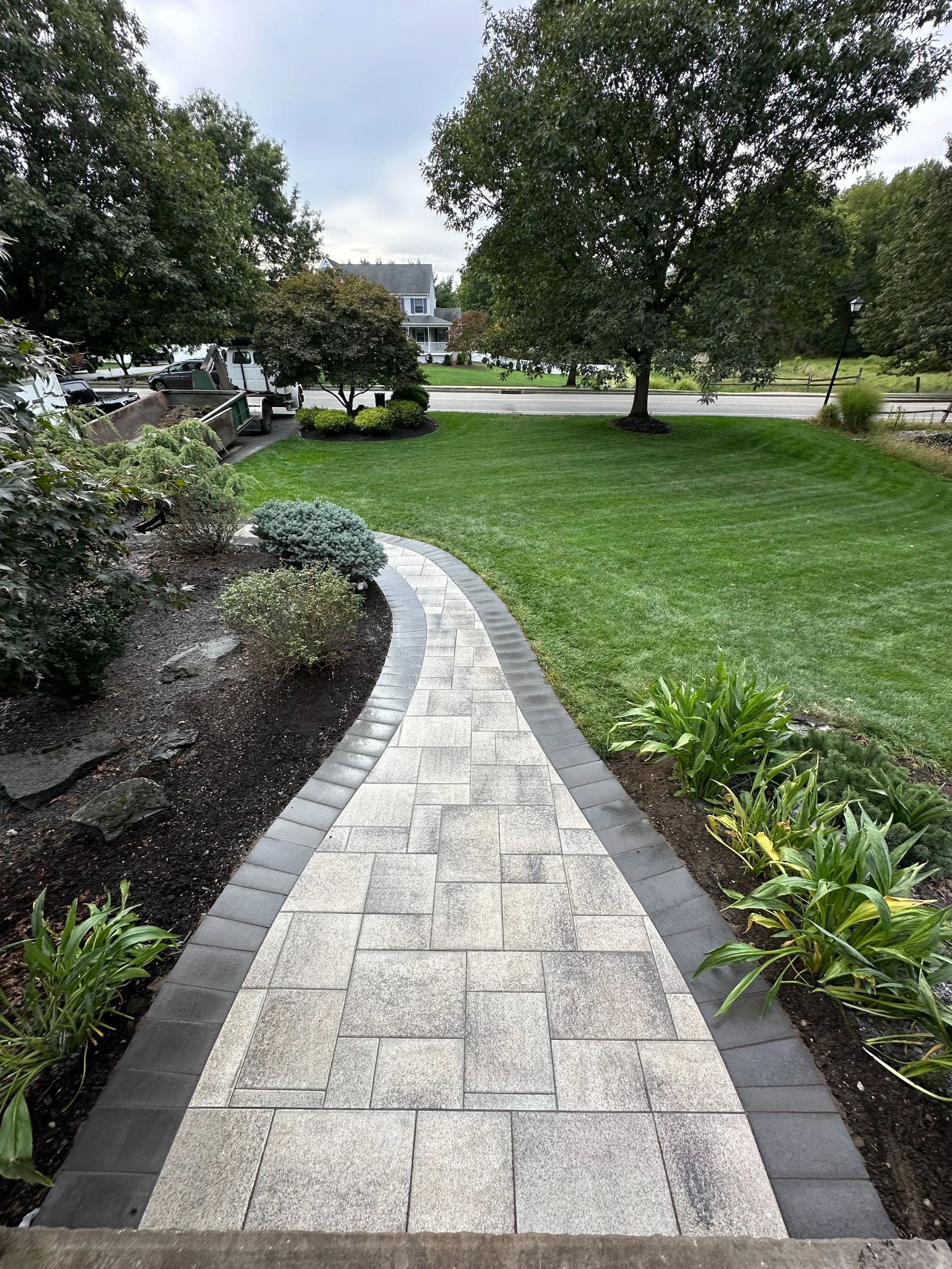 A paved walkway leading from a porch to a grassy yard with trees and bushes, and a street with houses in the background.