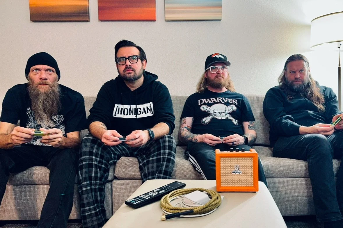 Four men sitting on a beige sofa in a living room with paintings above and a lamp on the side. They are holding gaming controllers, with an orange portable speaker, a remote control, and cables on the coffee table in front of them.