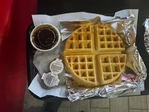 A breakfast tray with a waffle, a cup of syrup, and small containers of butter and syrup.