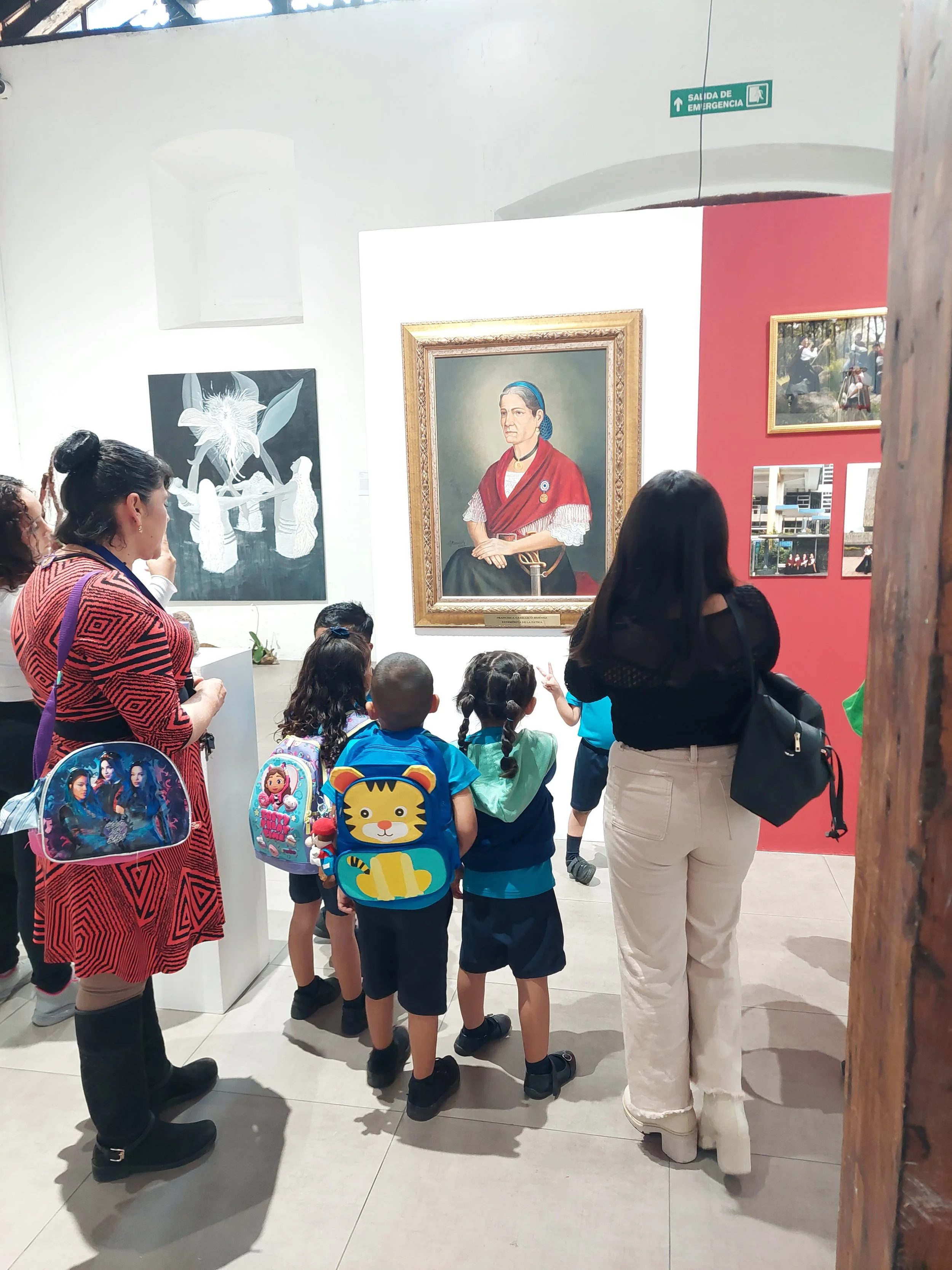 Group of children and adults gathered around a portrait of an older woman, in an art gallery.