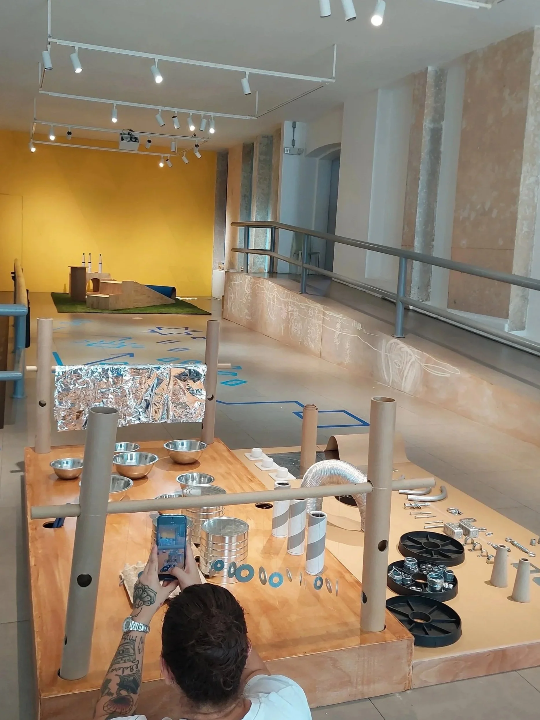 A person with tattoos on their arm is taking a photo with a smartphone of a workspace with various metallic and plastic parts, tubes, and wheels. There are wooden posts surrounding the workspace and a partly covered table. In the background there is a stage with a yellow wall and some small structures on a green mat, with spotlights on the ceiling.