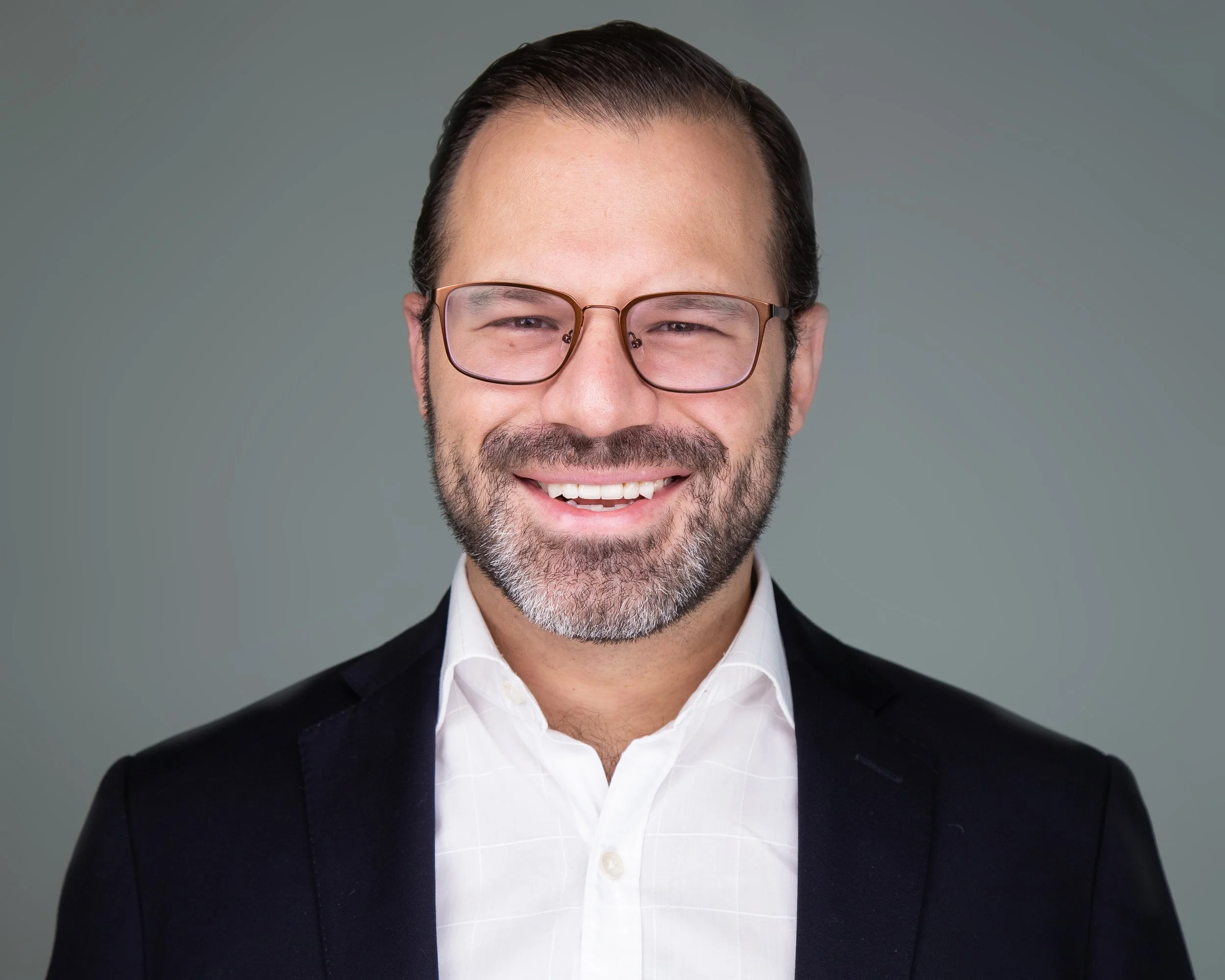 Headshot of smiling man with glasses, dark hair, and a beard, dressed in a white shirt and dark blazer against a plain background.