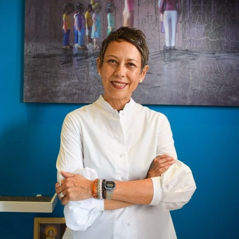 A woman with short dark hair smiling, wearing a white shirt with rolled-up sleeves, standing with her arms crossed in front of a blue wall and a large colorful artwork behind her.