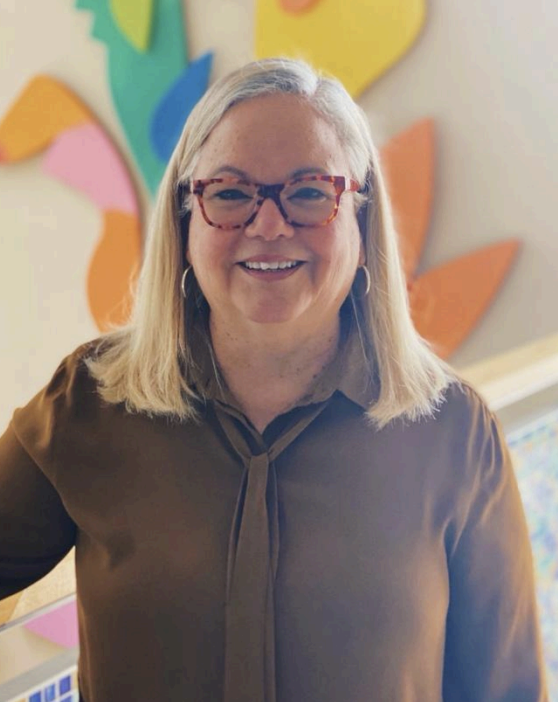 A smiling woman with blonde hair, glasses, and hoop earrings, standing in front of colorful paper heart decorations.