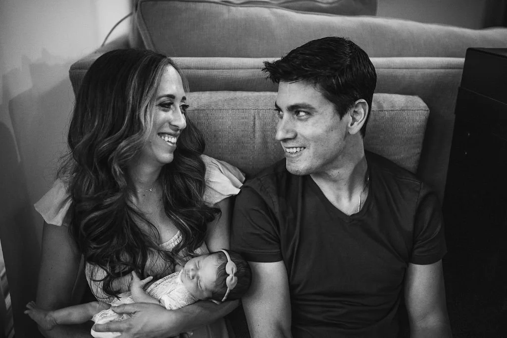 A woman with long, wavy hair holding a newborn baby, smiling at a man with short dark hair seated next to her; they are inside a room with a couch in the background.