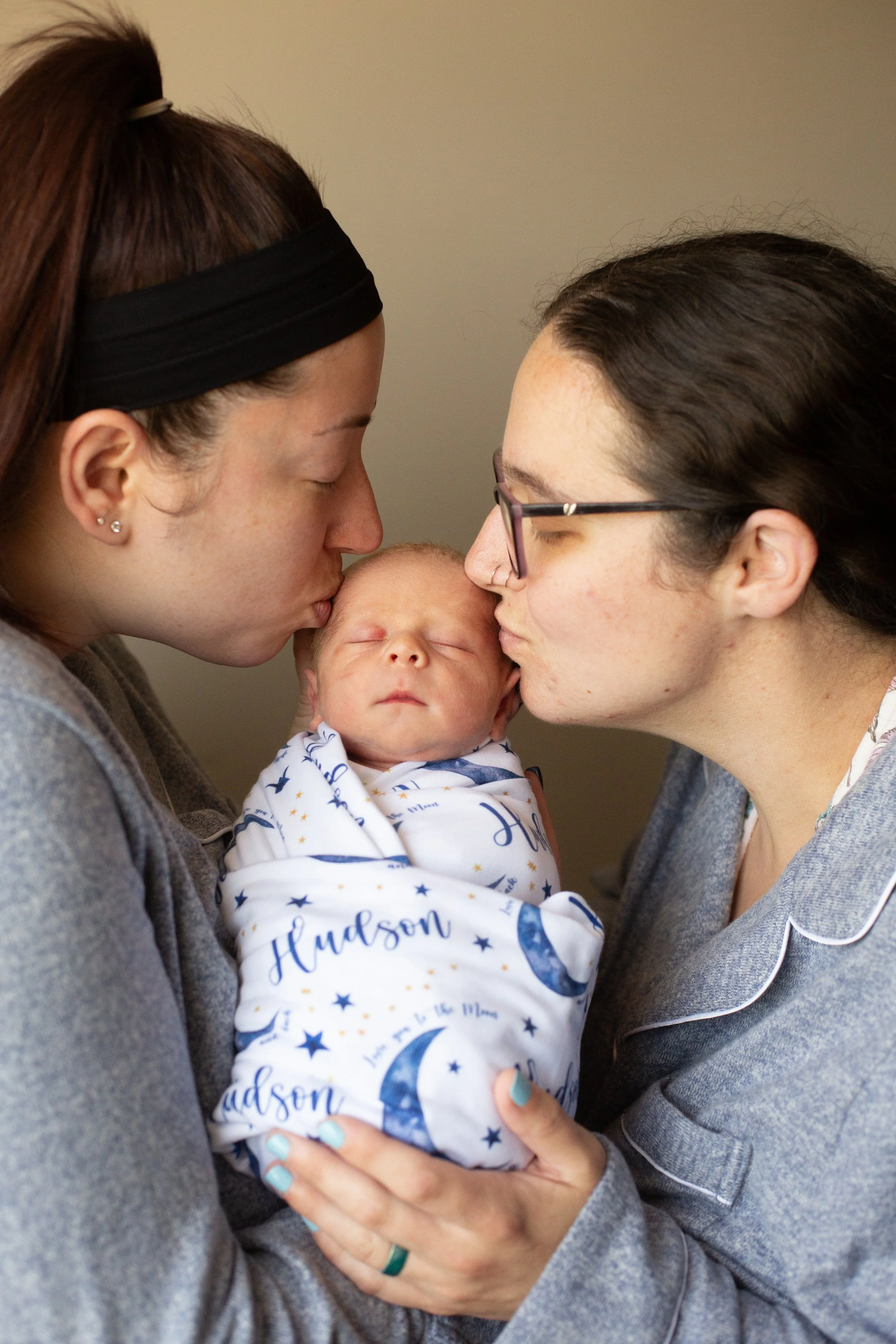Two women, one with glasses and one with a black headband, kissing a sleeping newborn wrapped in a blanket with moon and star patterns.