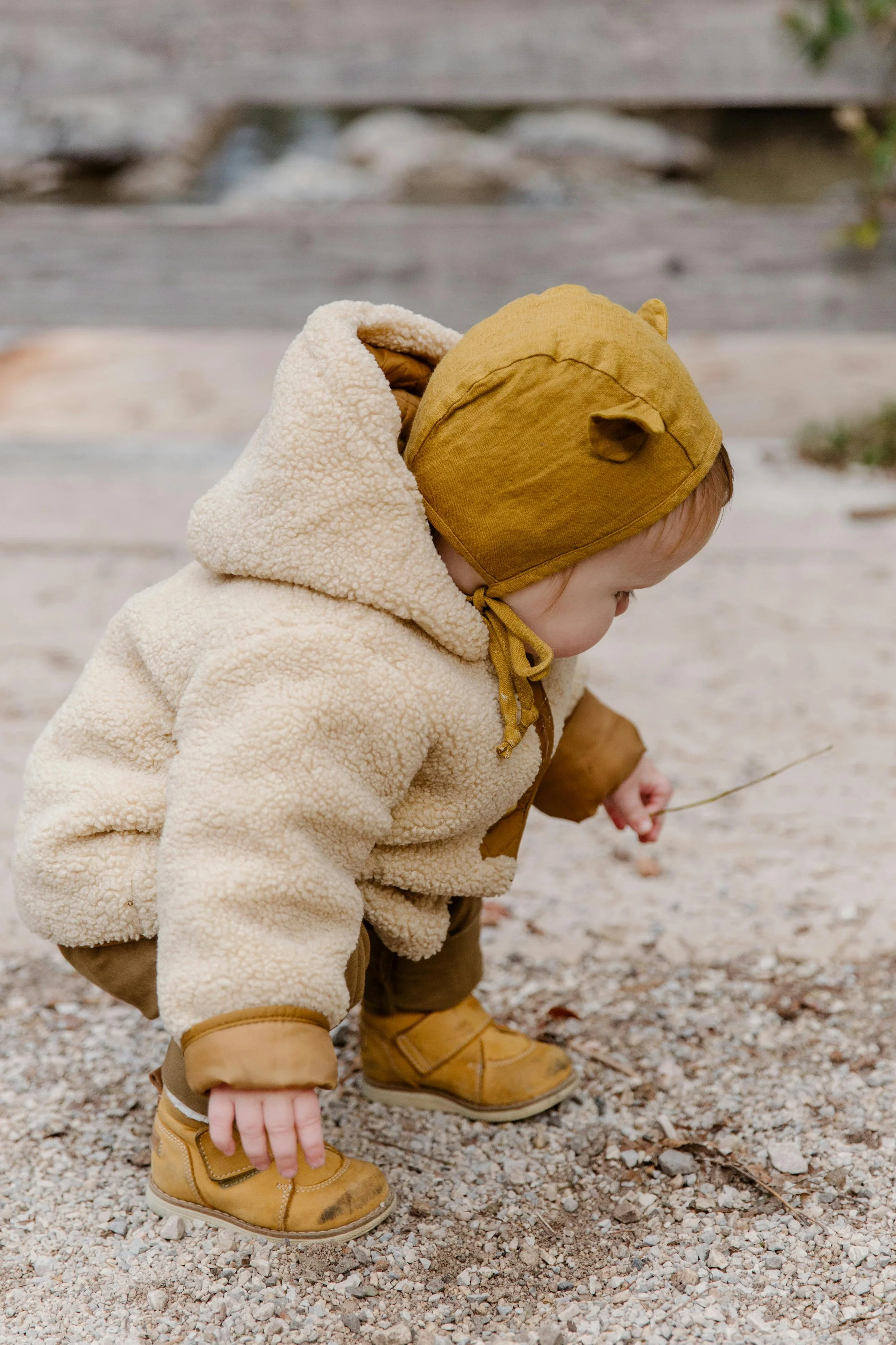 A young child wearing a beige fluffy jacket, yellow hat with bear ears, and yellow boots, crouching down and playing in the gravel outdoors.