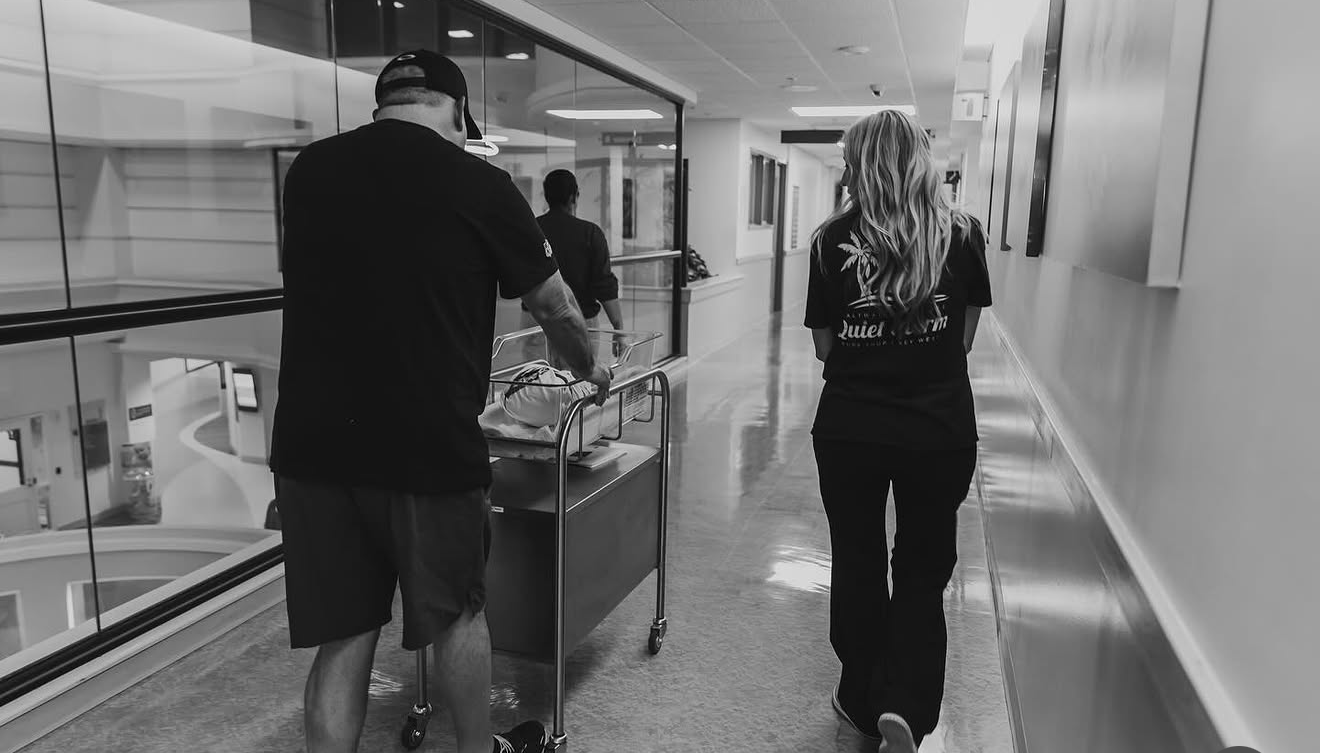 Hospital hallway with a woman and a man pushing a cart with an object on it, both wearing black clothing, with the woman walking ahead and the man behind, viewed from the back.