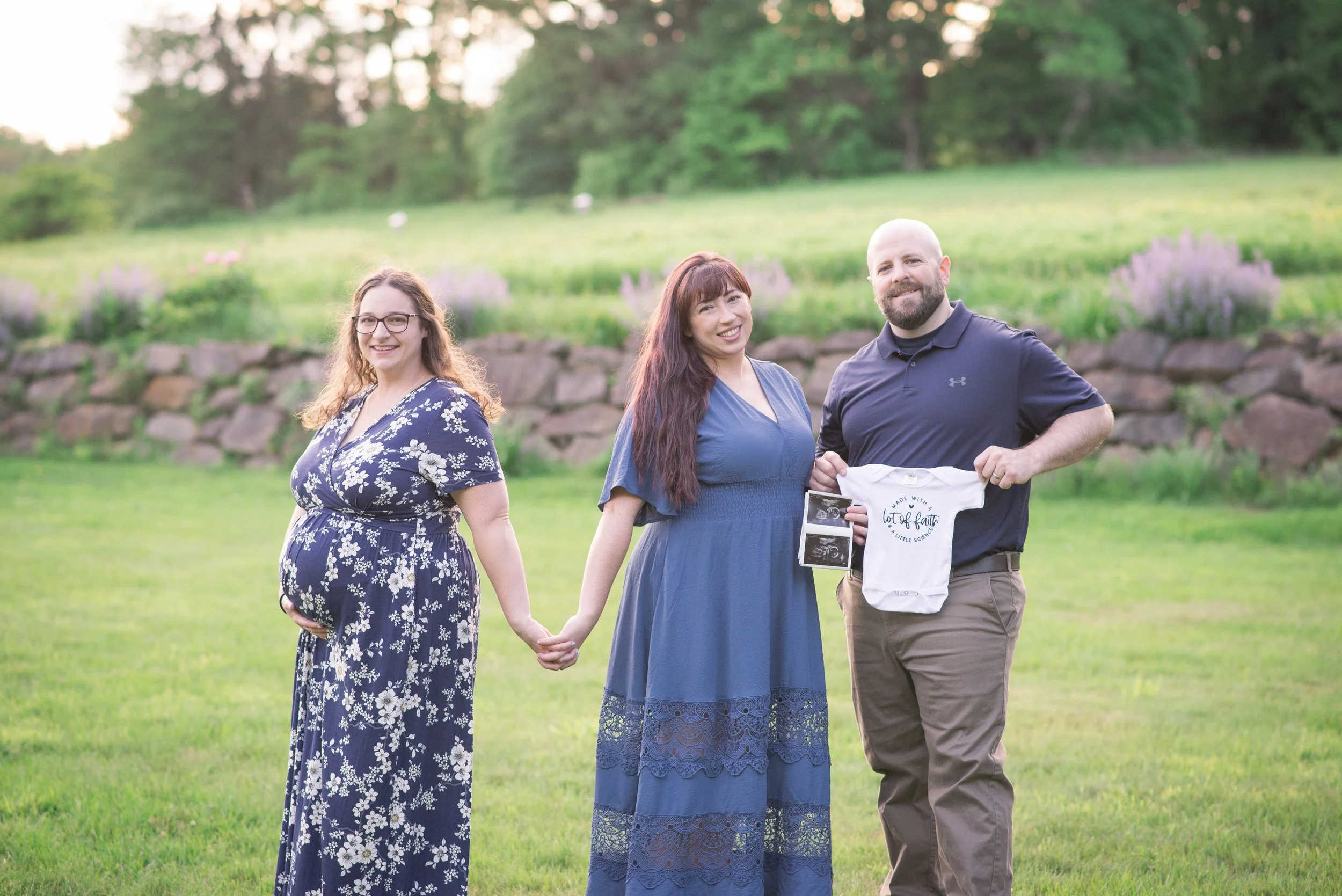 A happy pregnant woman holding hands with her partner, a man holding a baby onesie and ultrasound pictures, standing outdoors on a grassy field with a stone wall and trees in the background.