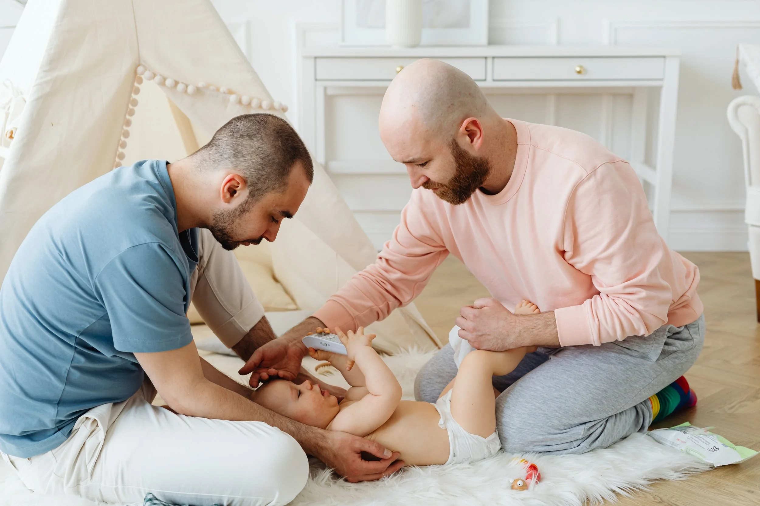 Two fathers care for their baby together on a soft blanket in a sunlit nursery