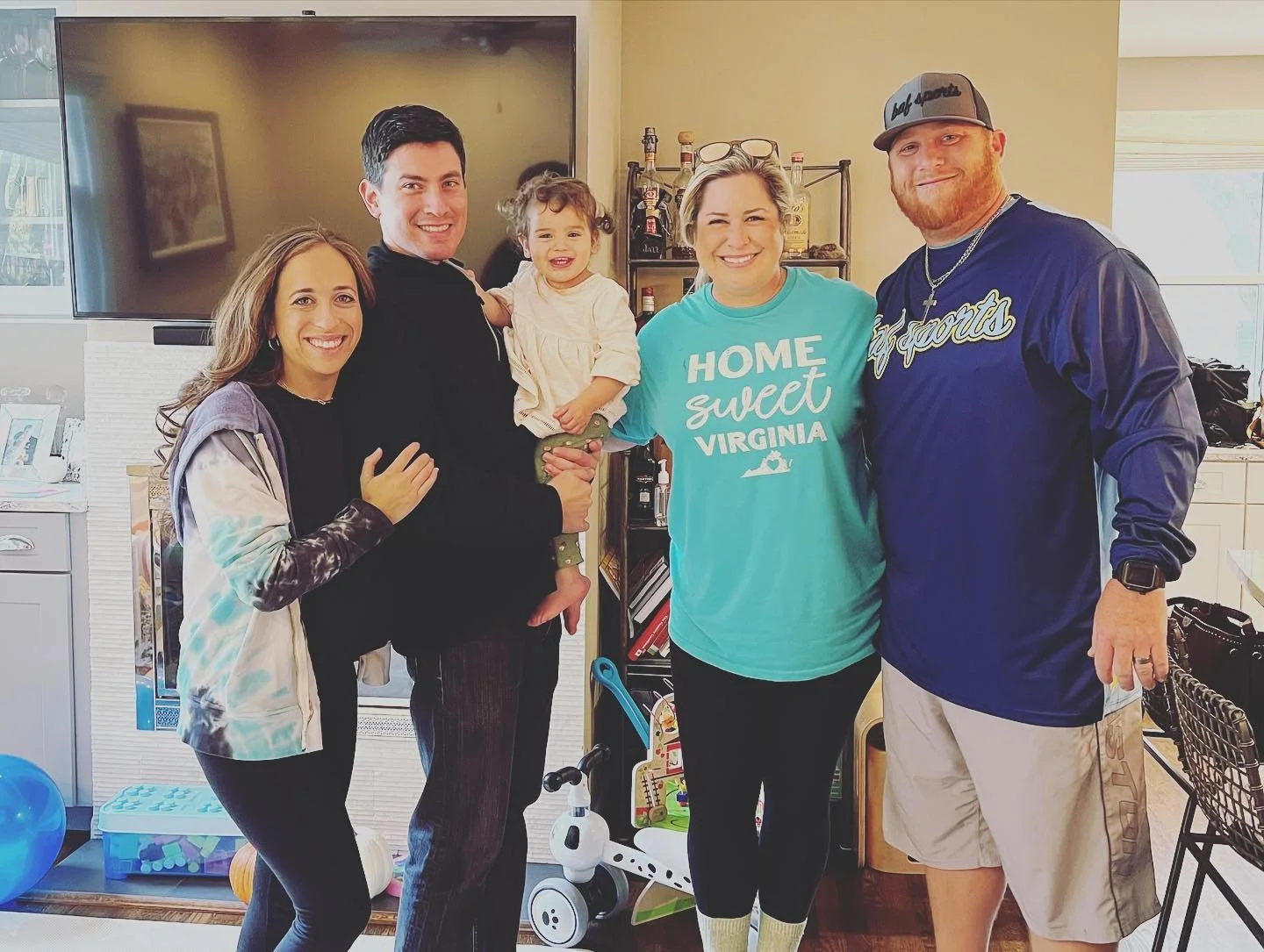 Group of five people, including a young girl, smiling in a cozy living room with a large TV and various household items, celebrating together.