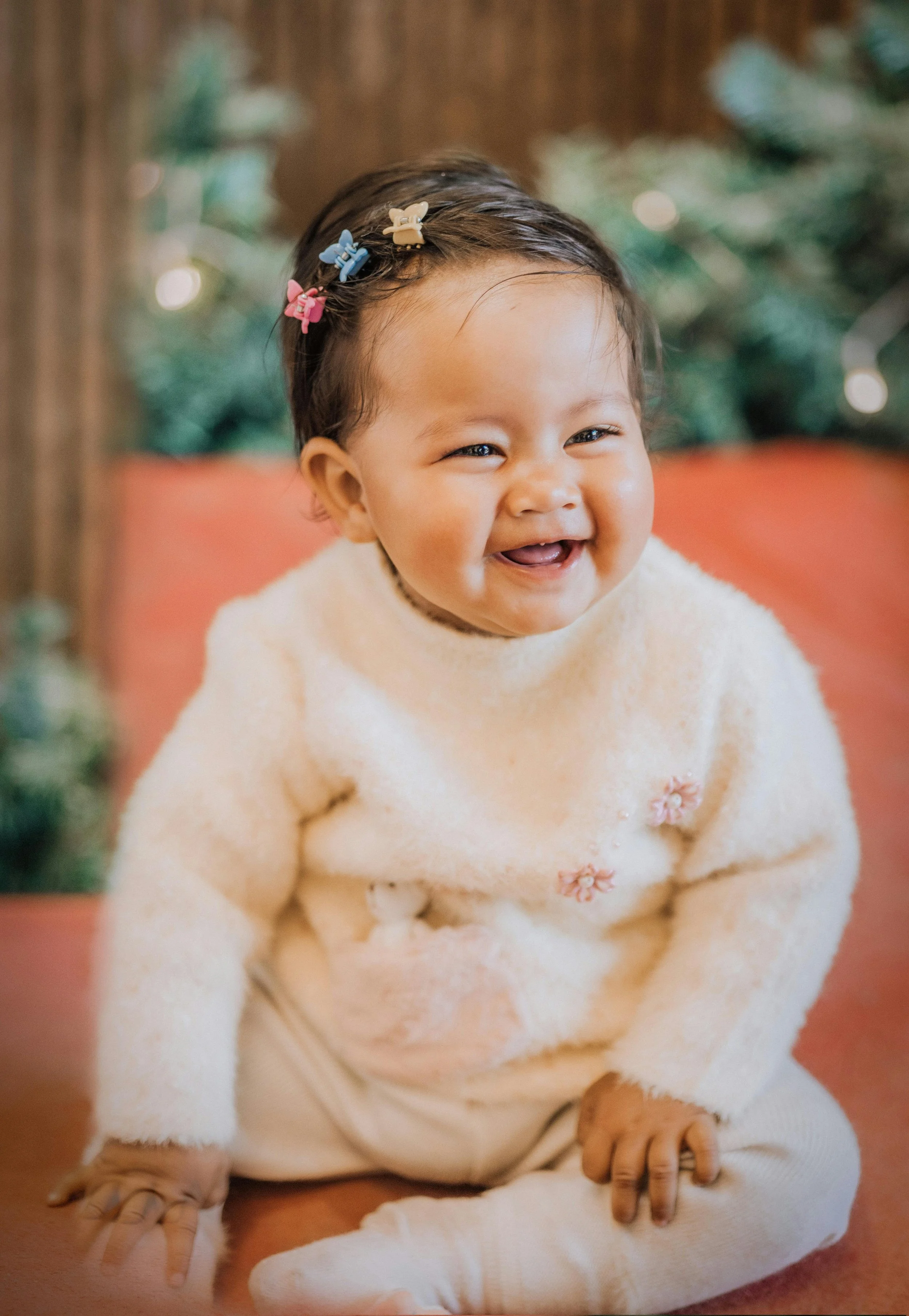 A smiling toddler girl with dark hair, wearing a cream-colored fuzzy sweater with pink flower embellishments and beige pants, sitting on a reddish surface with a blurred Christmas tree in the background.