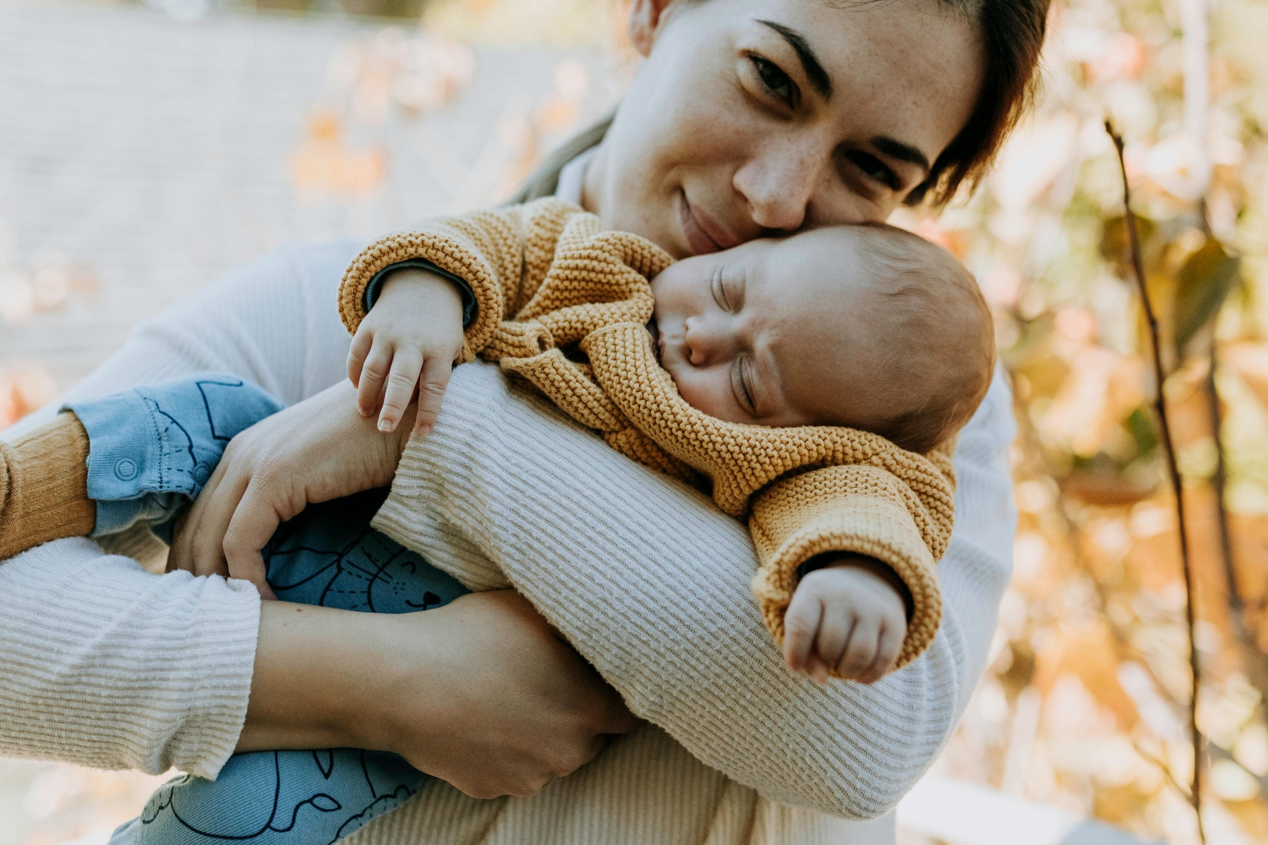 A woman holding a sleeping baby outdoors during autumn, surrounded by fall foliage.