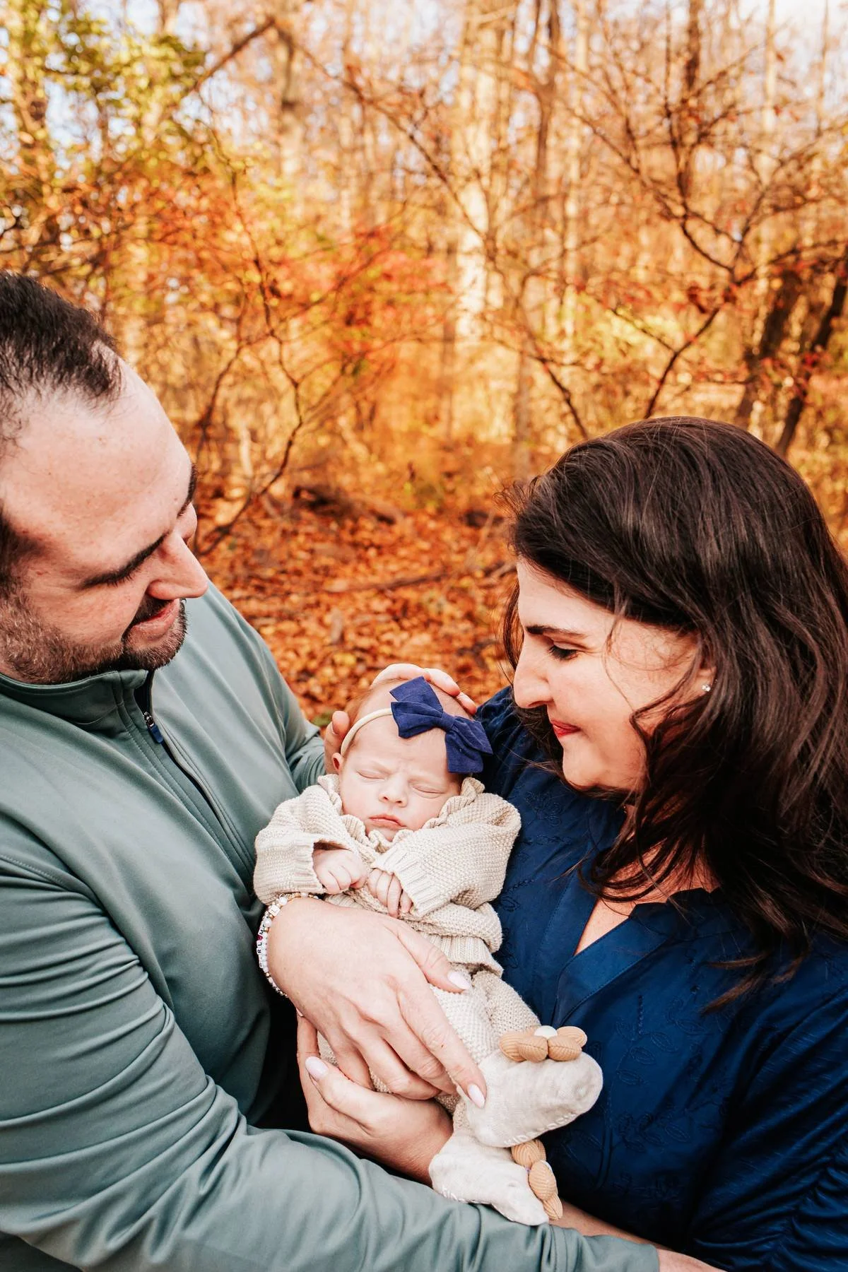 A family holding a newborn outside in fall, with autumn-colored trees in the background.