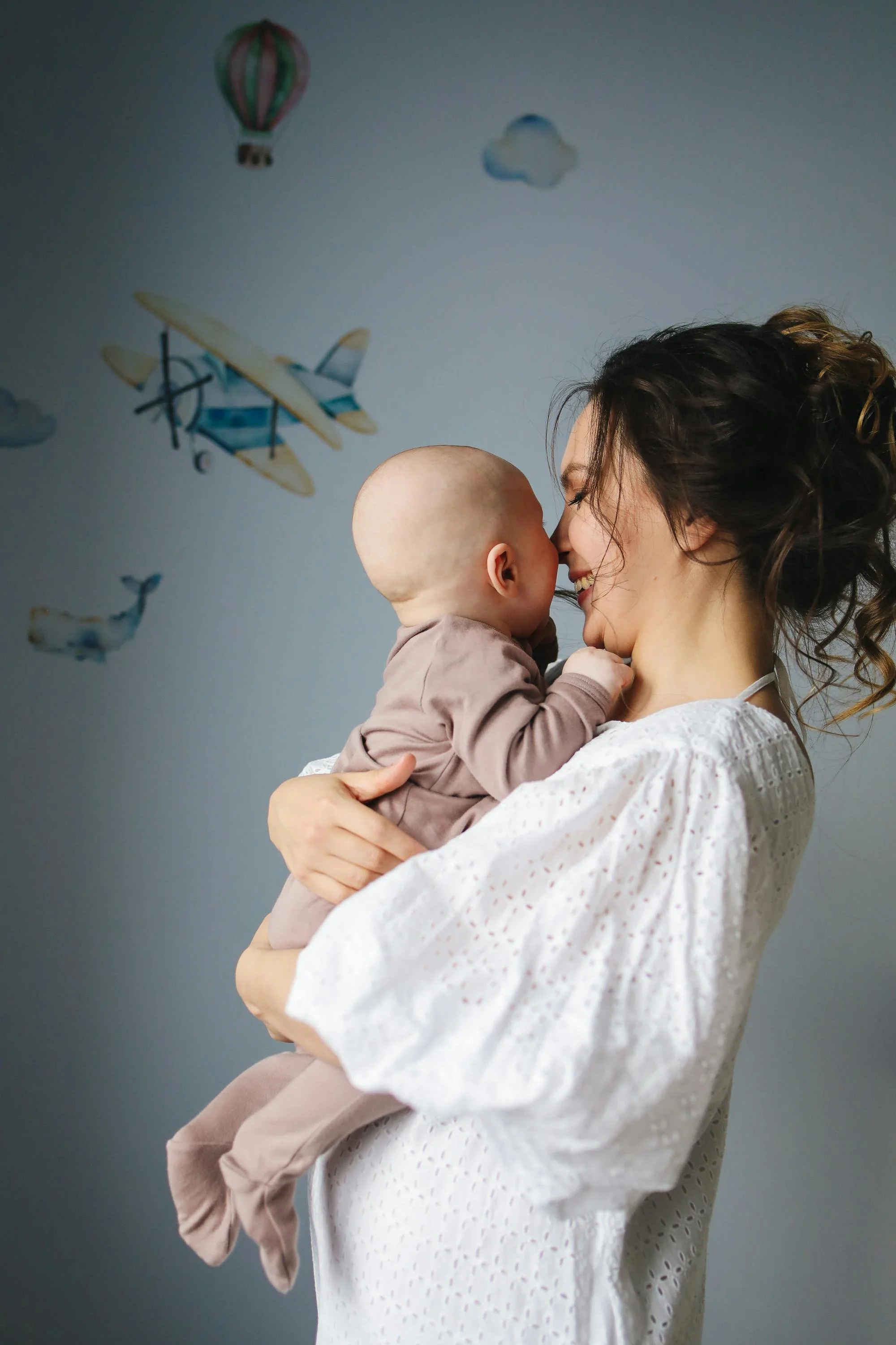 A woman with curly hair holding a baby with a bald head in front of a wall decorated with airplane and cloud stickers. They are touching noses and smiling.