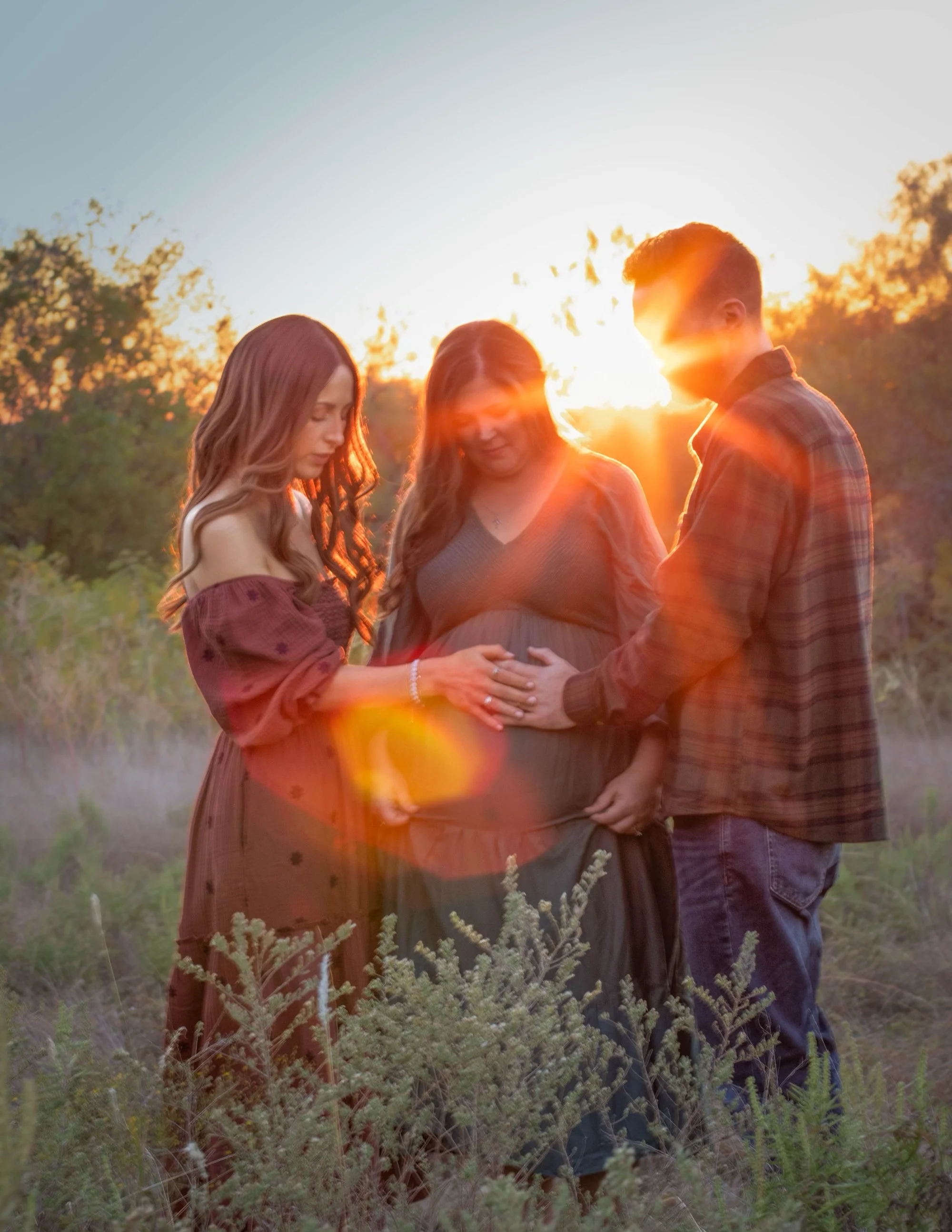 A pregnant woman with two others holding her belly outdoors during sunset.