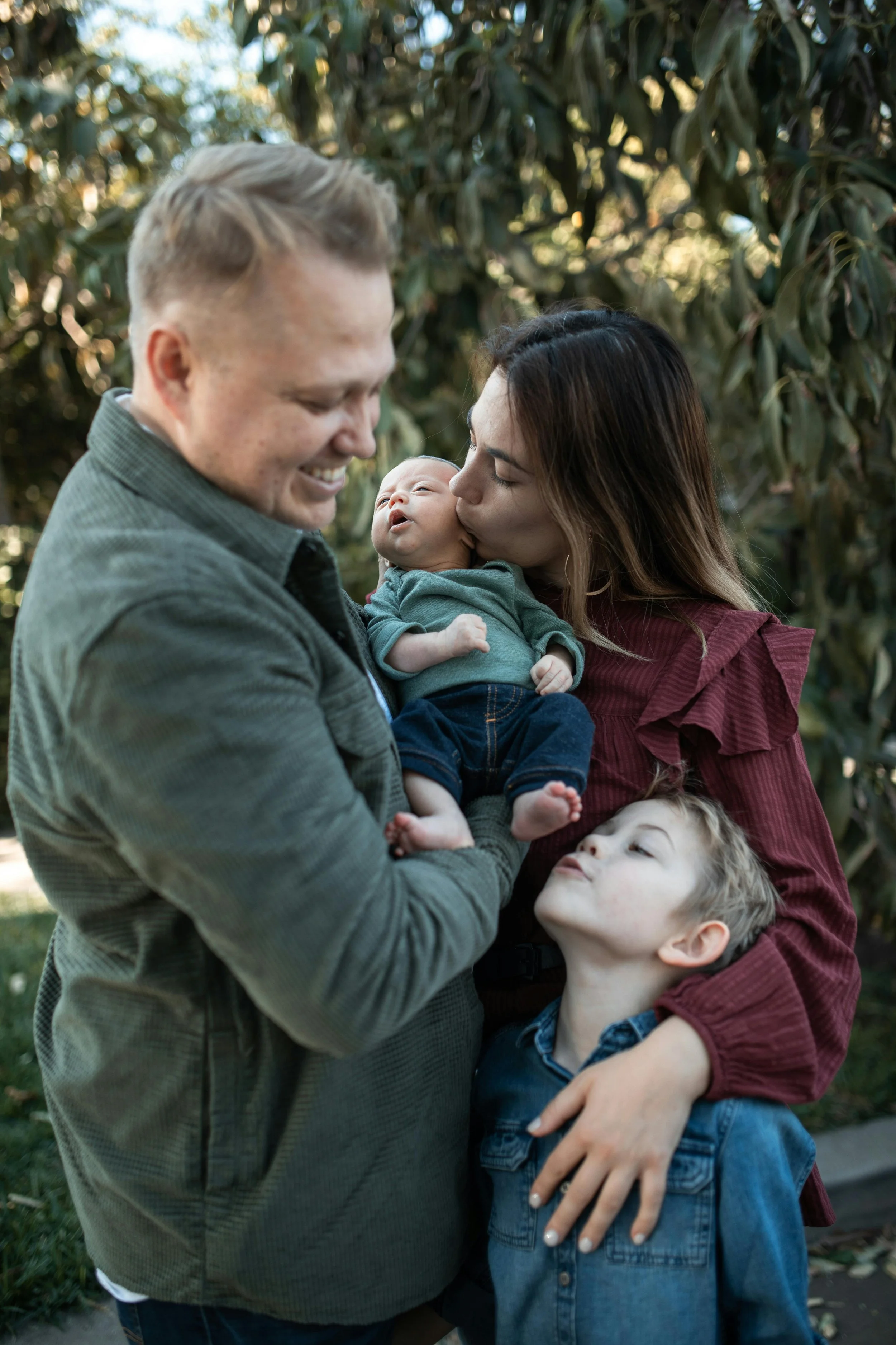 A family of four, including a man, woman, and two children, standing outdoors among trees. The woman is kissing a newborn on the head, and the man is smiling while holding the baby. The older child is standing nearby, looking up at the baby.