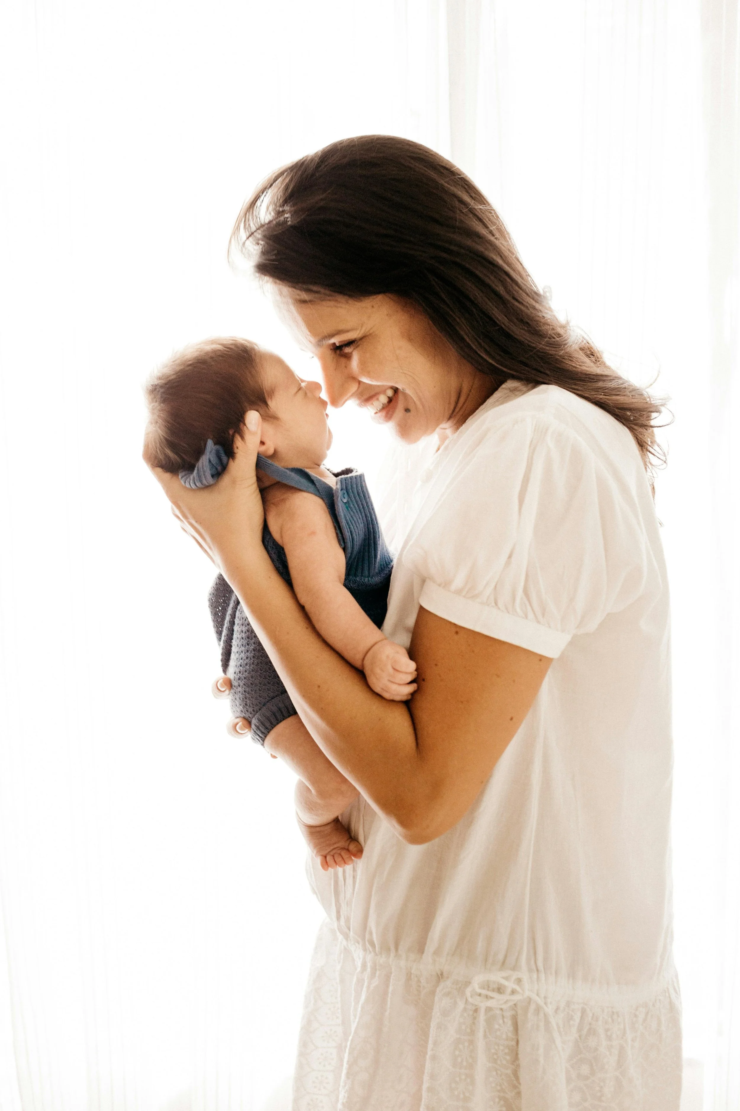 A woman smiling and holding a baby close to her face, both looking at each other affectionately in front of a bright window.