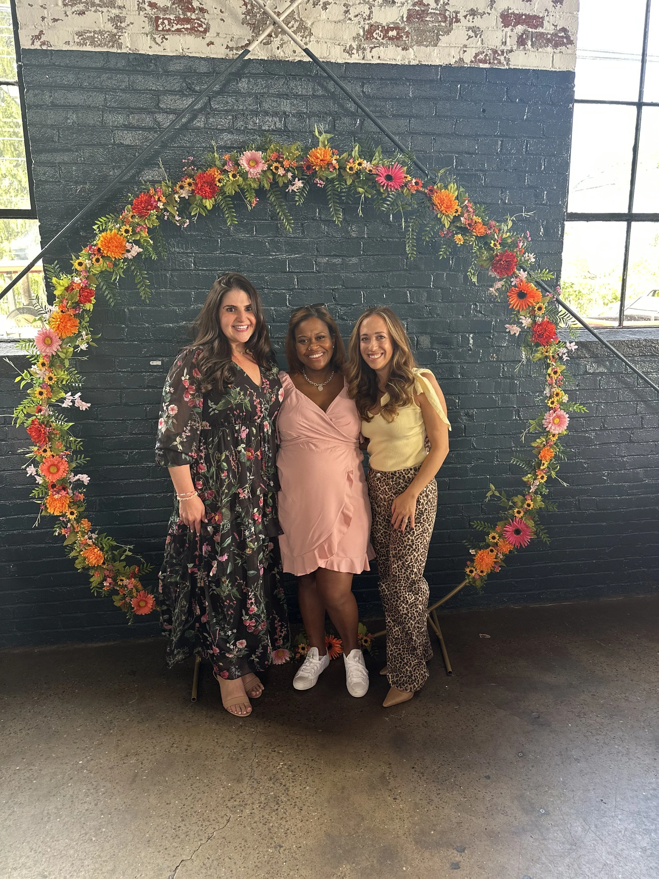 Three women standing together in front of a dark brick wall decorated with a large circular floral arch, all smiling.