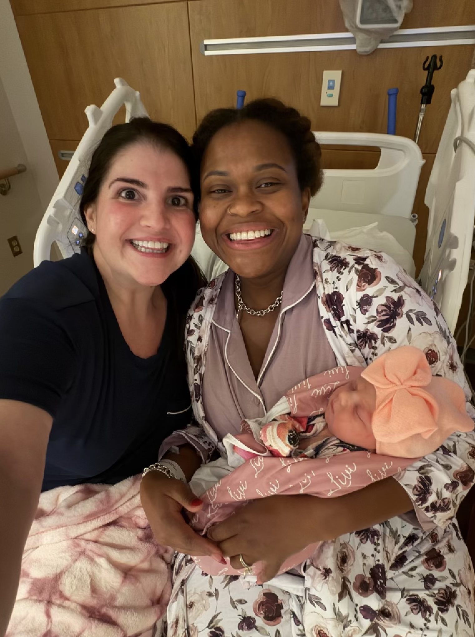 Two women smiling and taking a selfie in a hospital room. One woman, in pajamas, is holding a newborn baby wrapped in pink blanket with a pink bow hat. The woman in pajamas has dark skin and curly hair, the other woman has light skin and dark straight hair. Hospital bed and medical equipment are in the background.