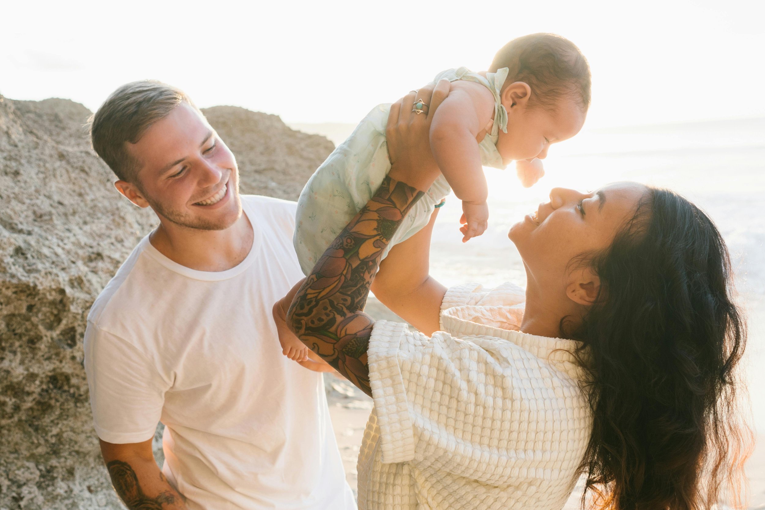 A family at the beach, with a woman holding a baby up while a man stands next to them, all smiling and enjoying the moment.