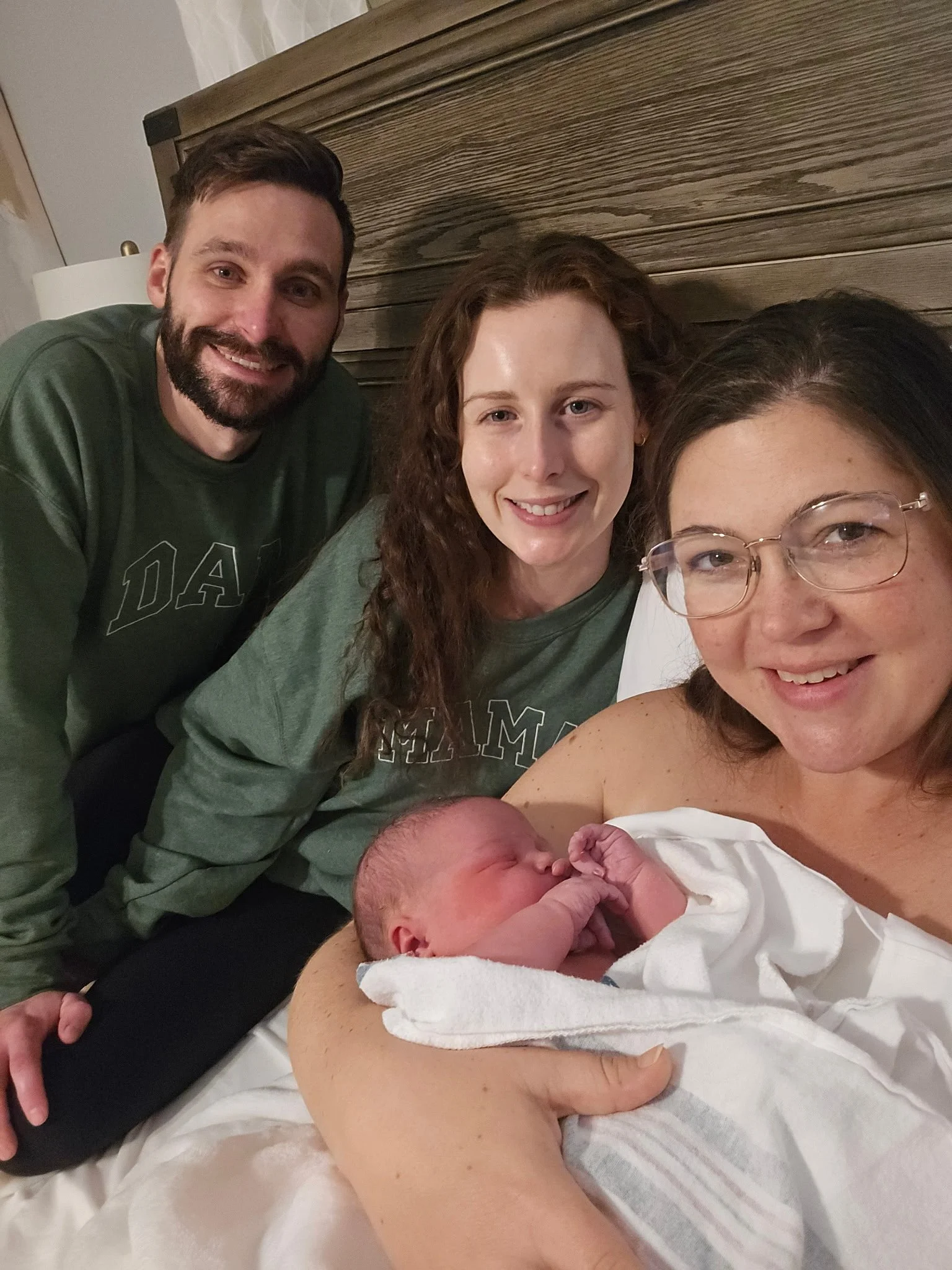 A woman holding a newborn baby in bed with three other adults around her, all smiling. The woman has glasses and the others appear to be the baby's family. The background has a wooden headboard and a lamp.