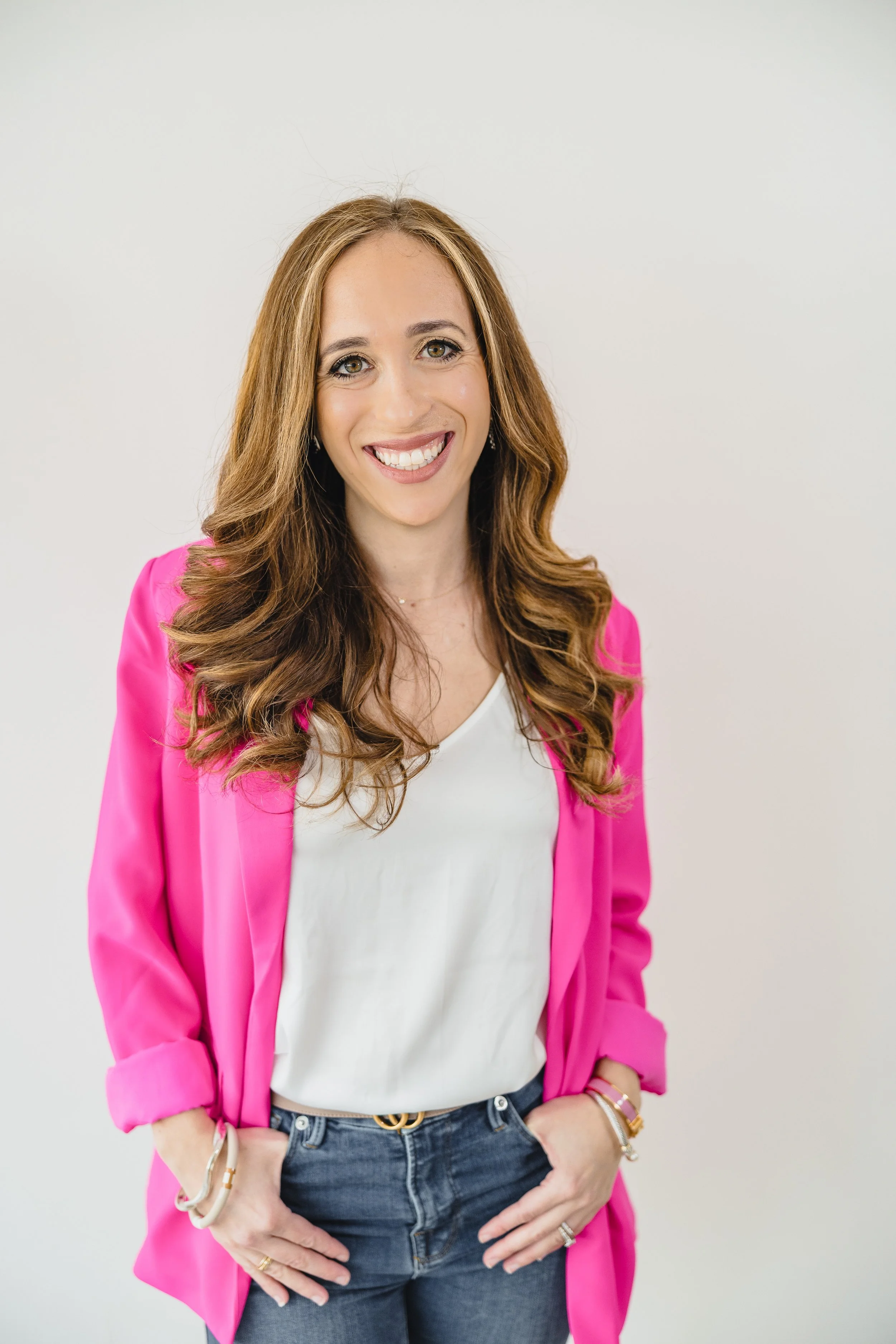 A woman with long wavy brown hair, smiling, wearing a pink blazer over a white top, blue jeans, and accessorized with bracelets and rings, standing against a plain white background.