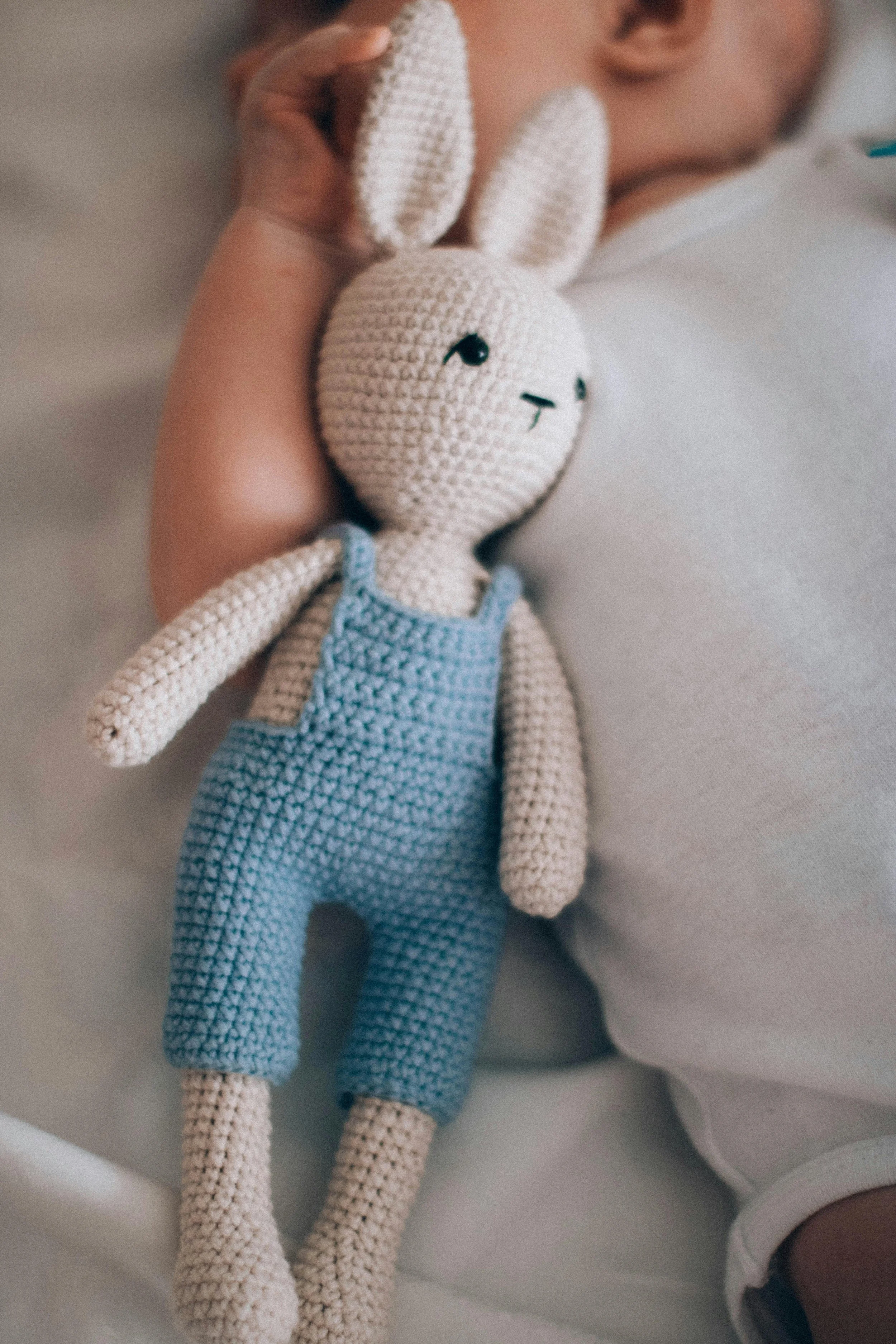 A hand holding a crocheted bunny stuffed animal with a blue outfit, placed on a person's shoulder.