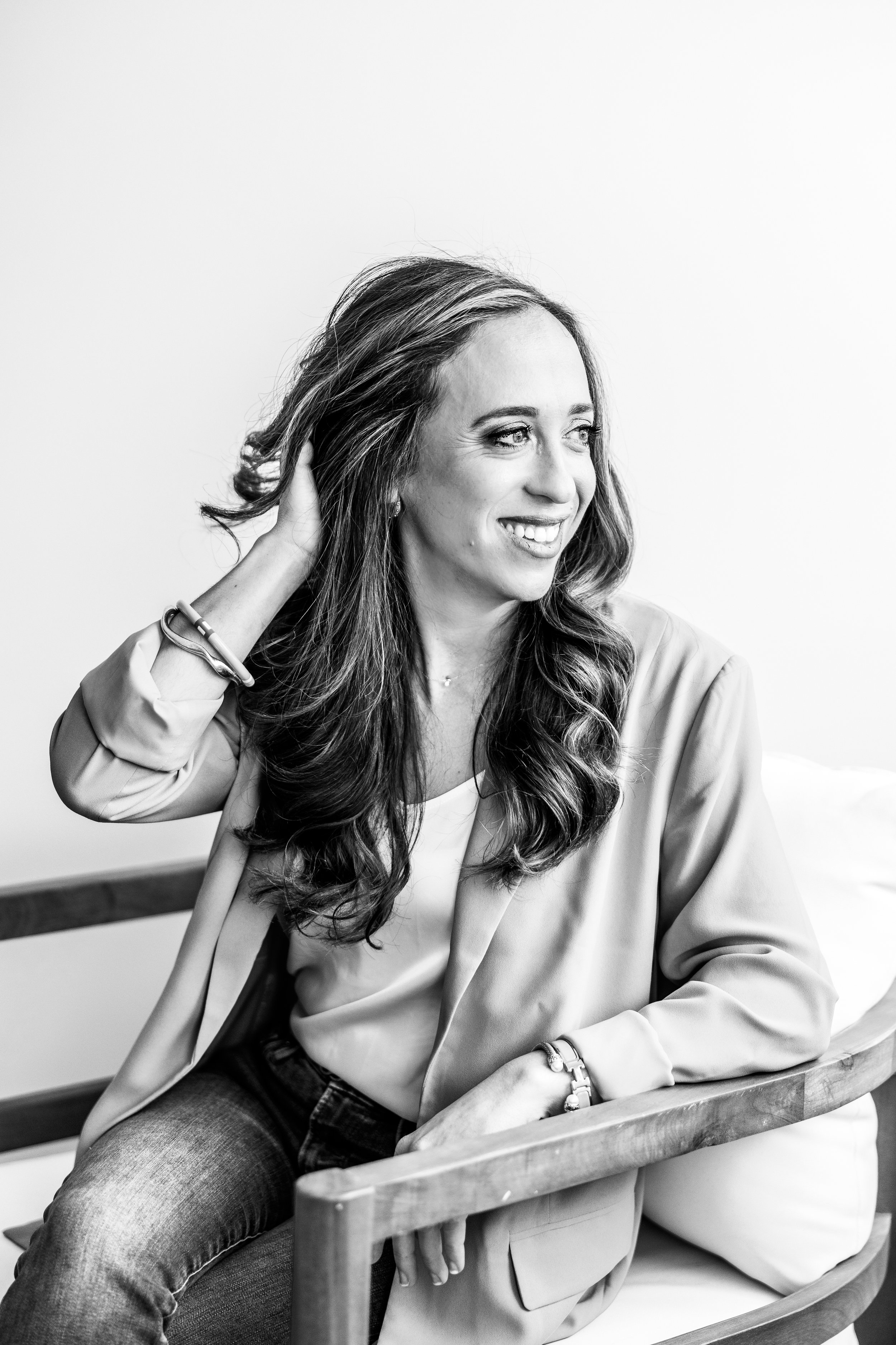 A woman with long wavy hair smiling and sitting on a wooden bench, wearing a blazer, a necklace, and bracelets, against a plain white background.