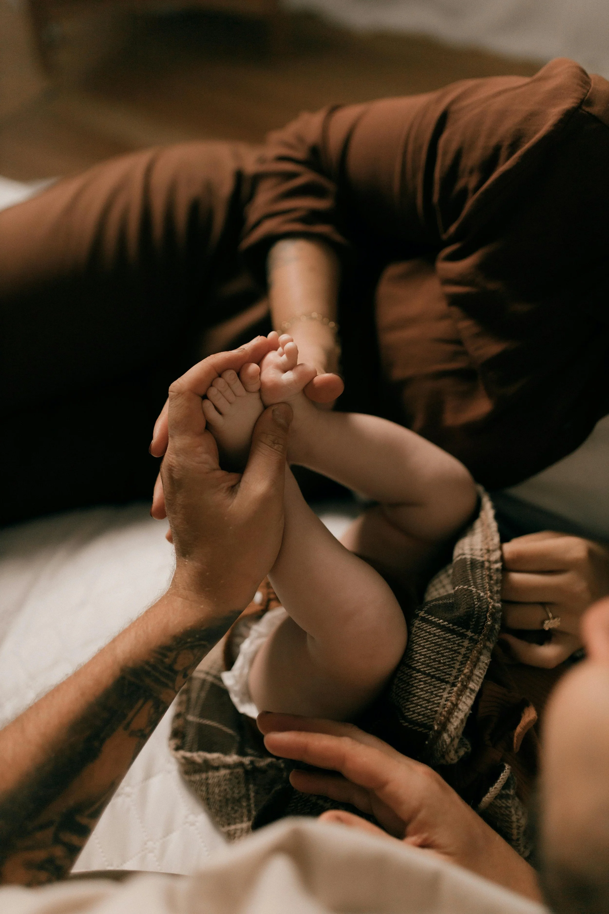 Person lying down holding a baby's foot, with a hand gently touching the baby's leg, in a cozy indoor setting.