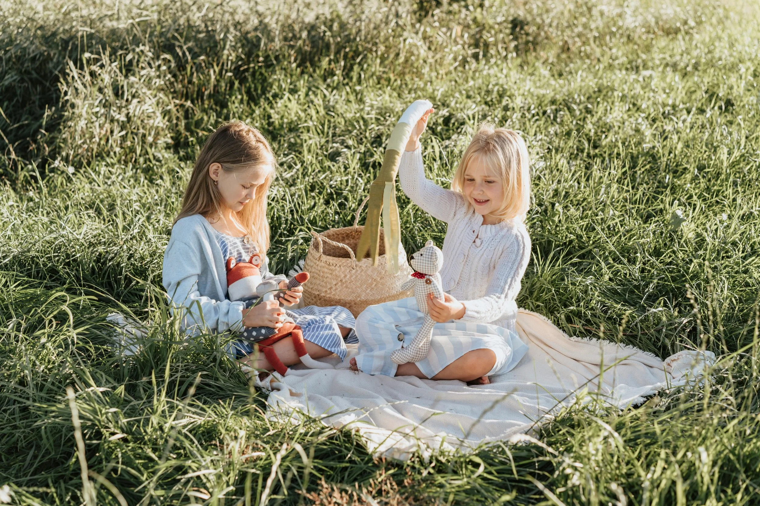 Two young girls sitting on a blanket in a grassy field, playing with stuffed animals and enjoying a picnic.