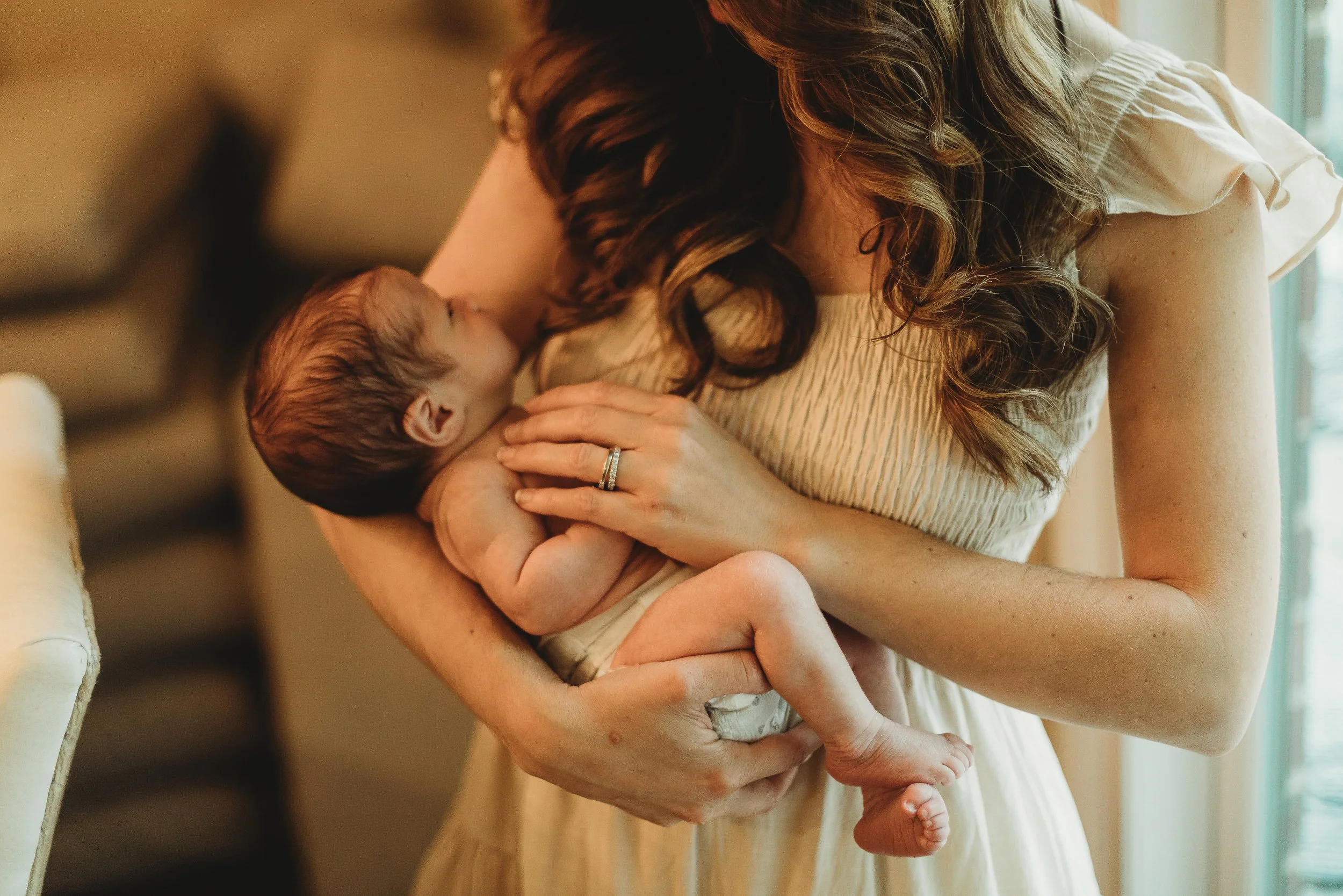 A woman with long wavy hair in a cream-colored dress is holding a newborn baby close to her chest, with the baby nursing. The focus is on the woman and baby, with soft lighting and a blurred background.
