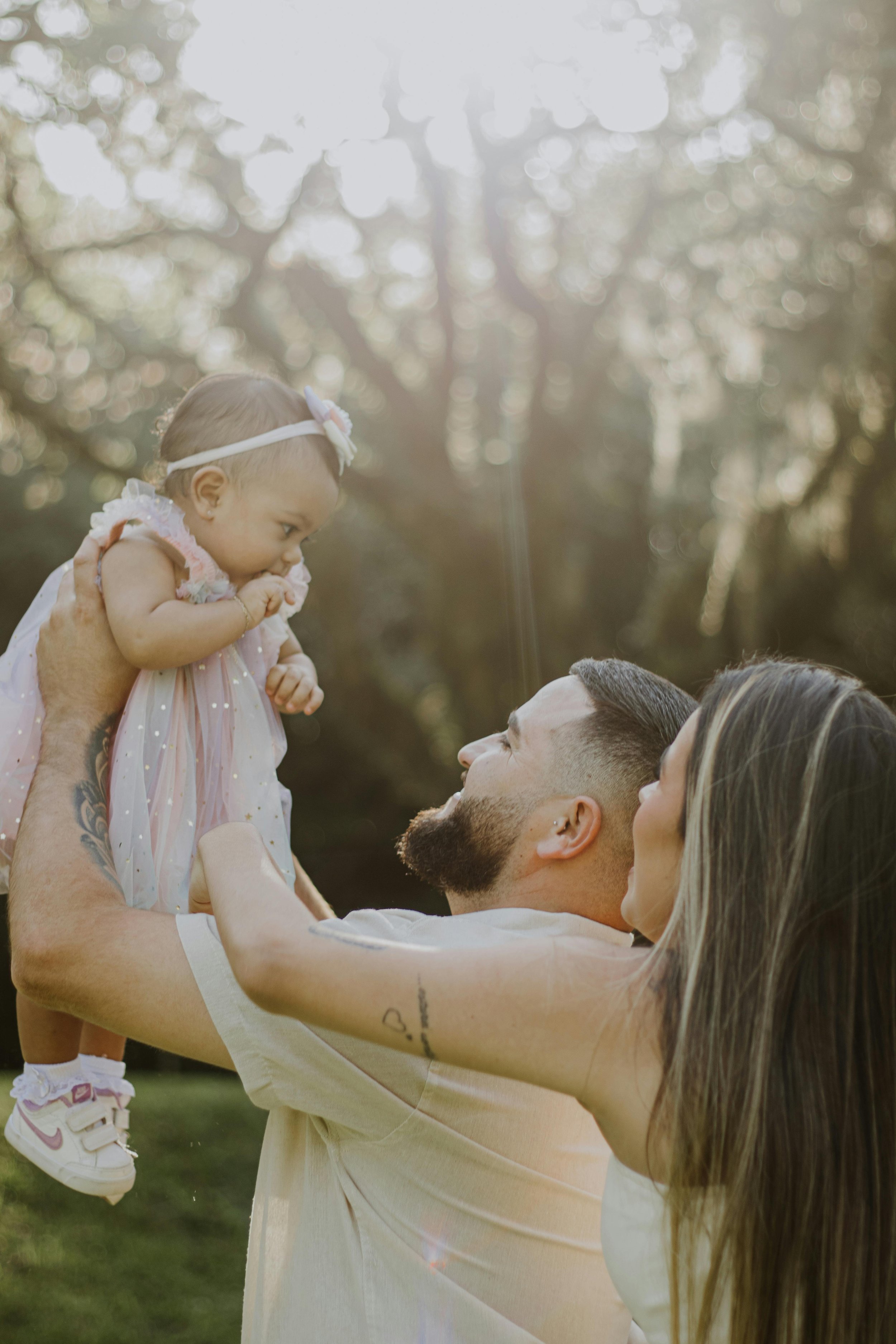 A man and a woman lift a young girl into the air outdoors on a sunny day with trees in the background.
