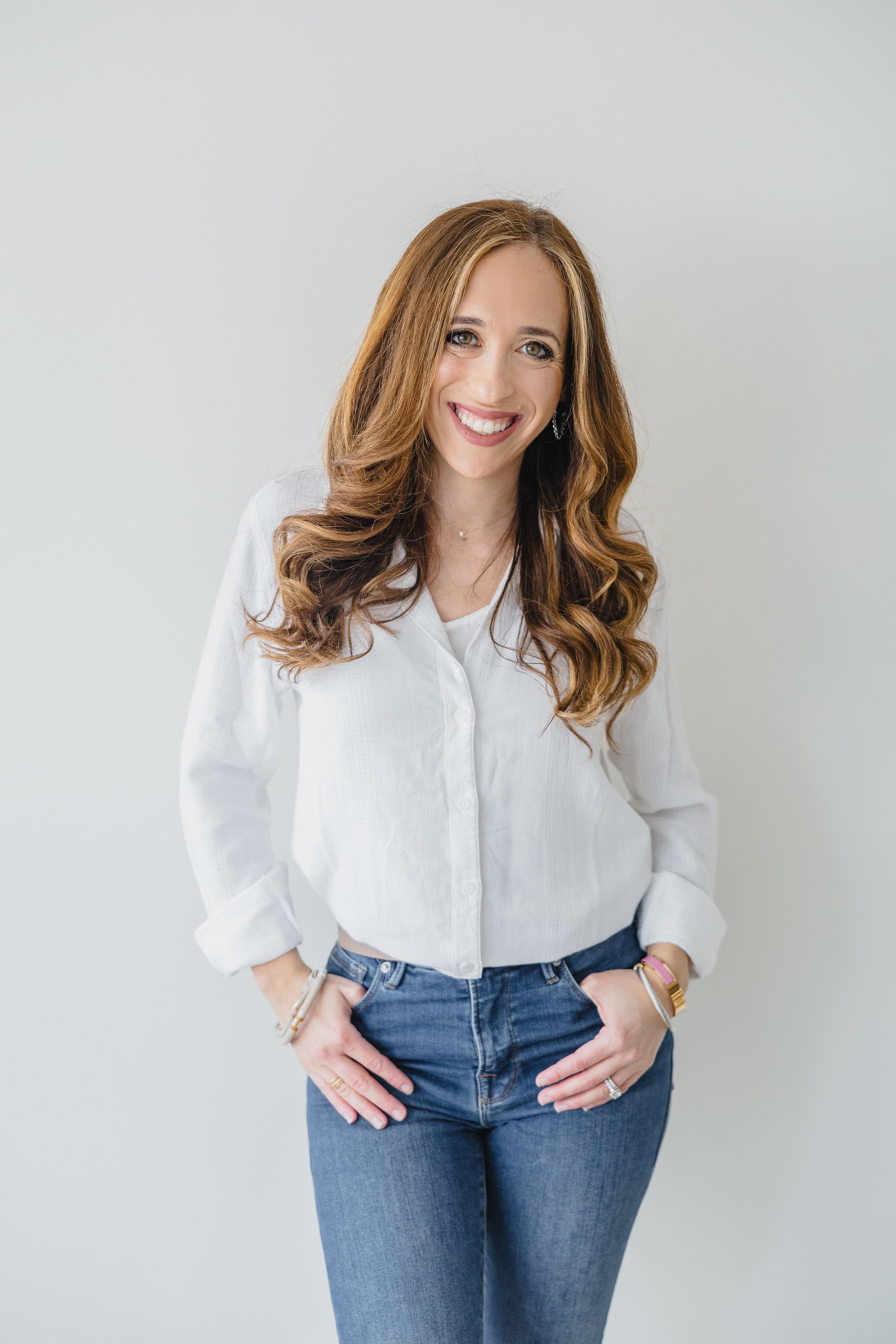 A woman with long, wavy, light brown hair, wearing a white button-up shirt and blue jeans, standing against a plain white wall, smiling, with her hands in her pockets.