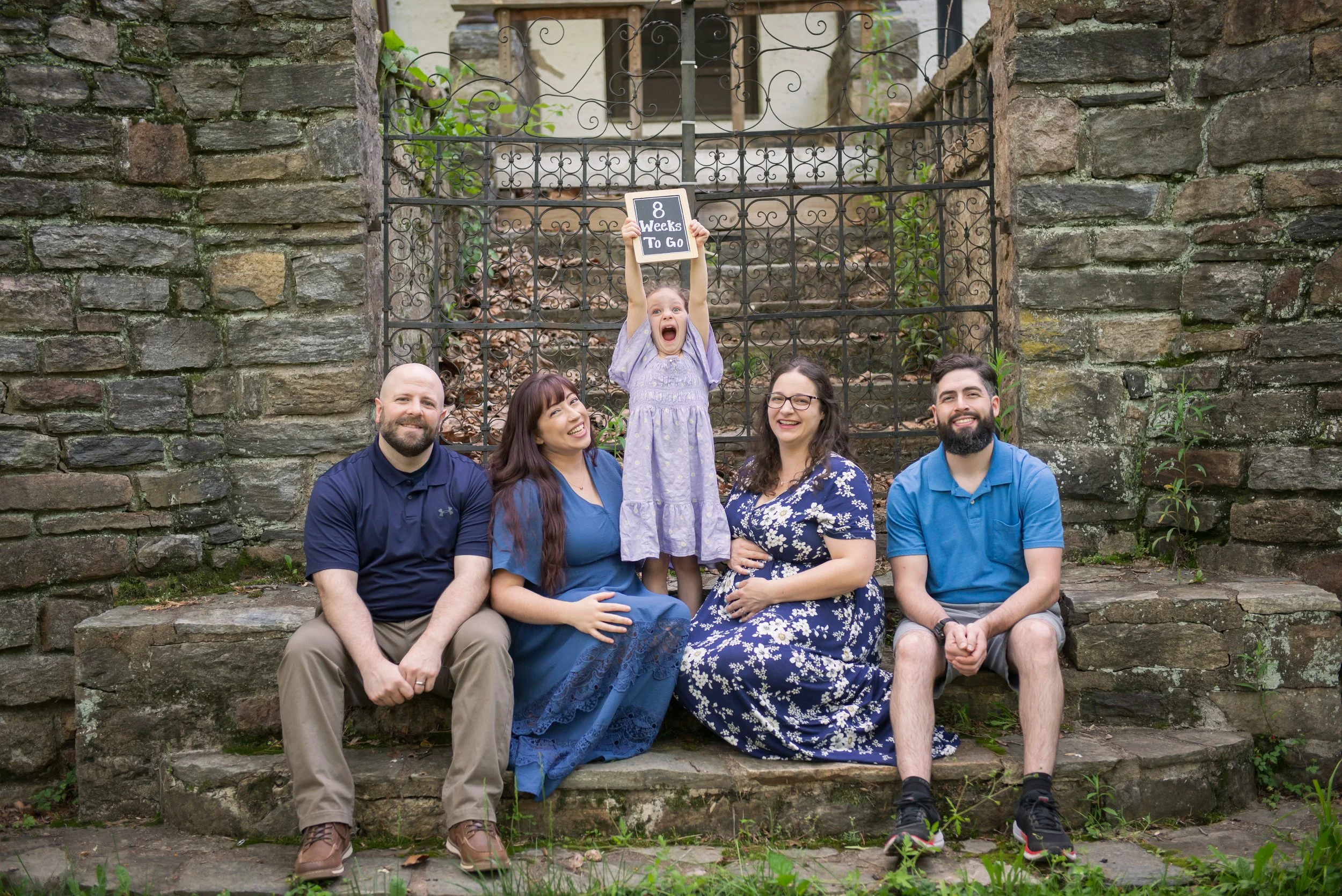 A family of five sitting on stone steps in front of a decorative wrought iron gate and stone wall, with a young girl standing between her parents holding a sign that reads '8 Weeks To Go', all smiling and looking happy.