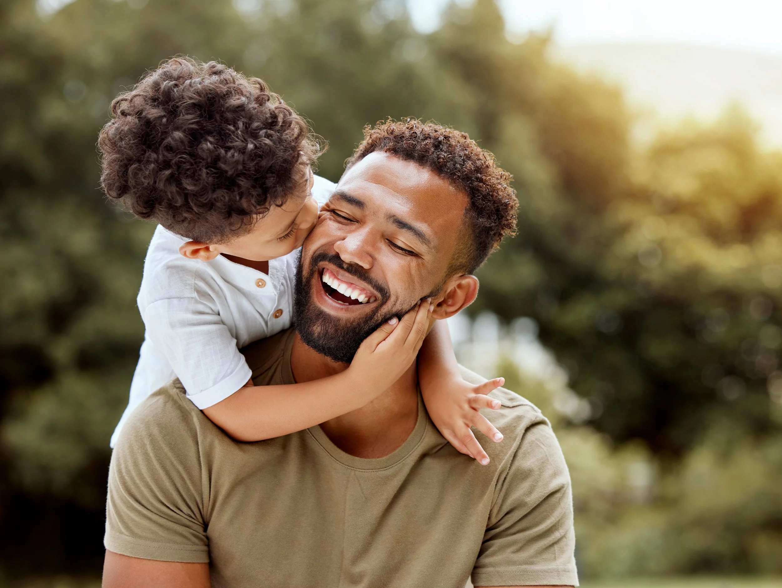 A father and son sharing a joyful moment outdoors, with the son kissing the father on the cheek, both smiling and surrounded by greenery.
